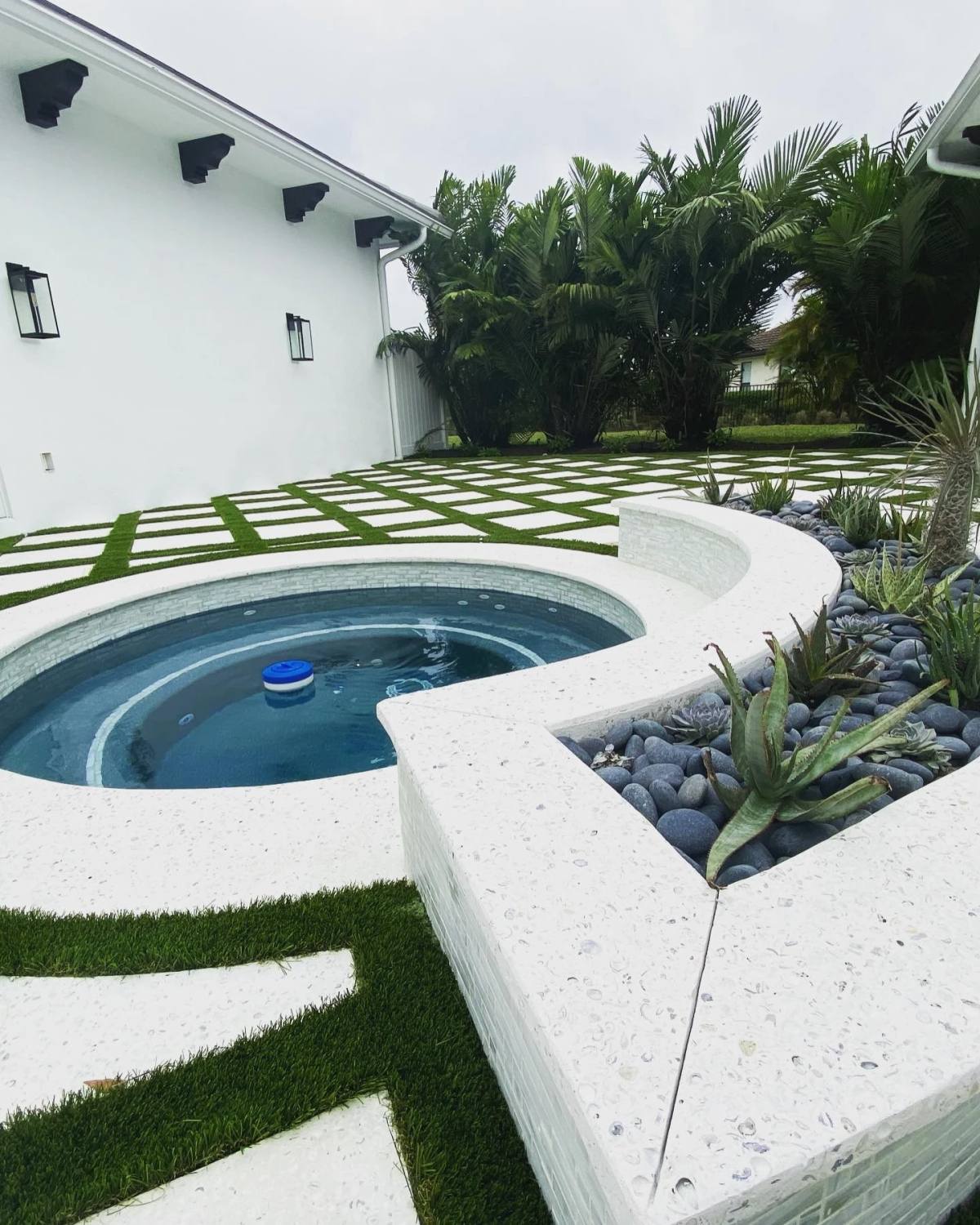 A curved pale surround and raised planter wrapping a round spa, with a checkerboard of square slabs set into artificial turf running beyond