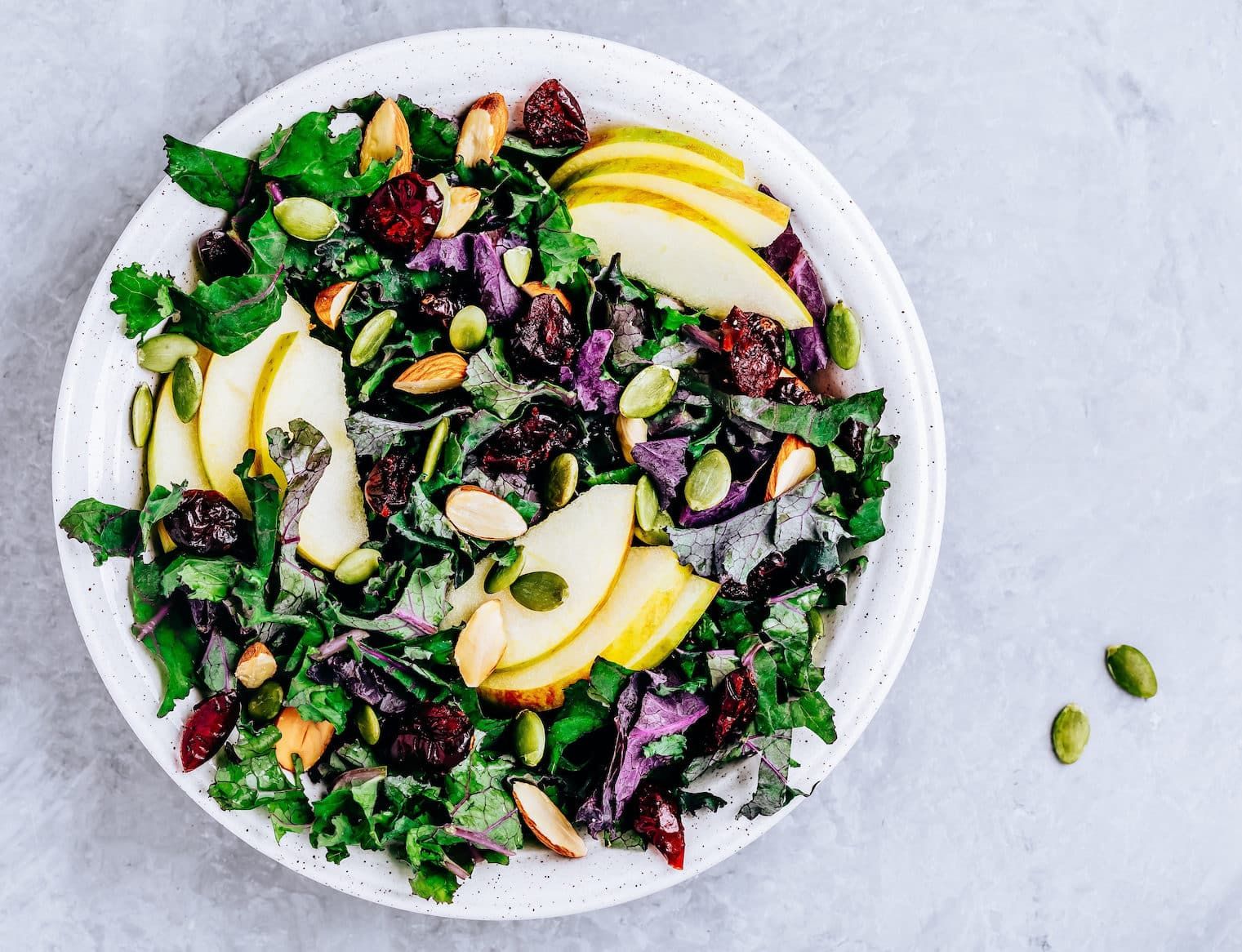 Hearty Kale with Crisp Apples, Spiced Pepitas and Goat Cheese in a white bowl on a grey table top