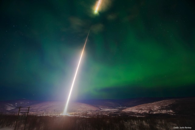 A bright-white trail marks a rocket's path as it ascends into a green aurora borealis in the night sky.