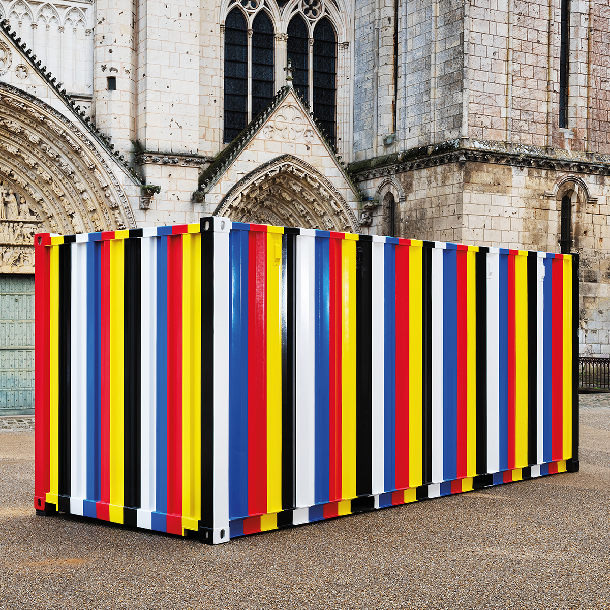 Shipping container painted in white, blue, red, yellow and black stripes, in front of cathedral