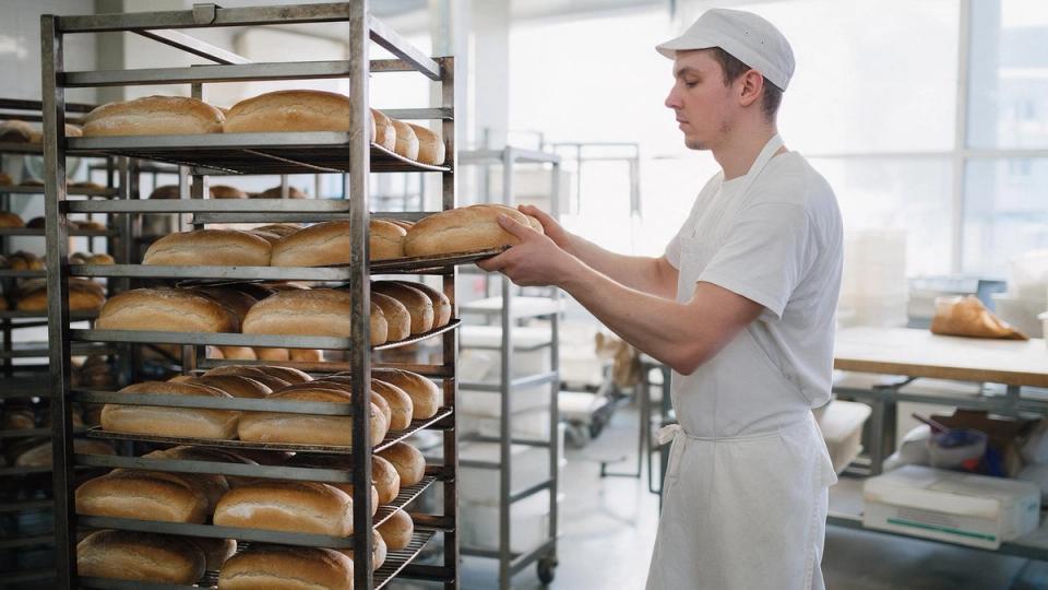 Bakery worker in white cap and apron loads a tray of fresh loaves onto a tall steel rack