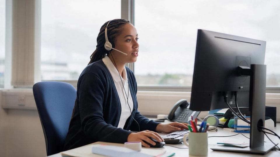 Telesales agent with braided hair talks through a headset while working at a desk by a large office window