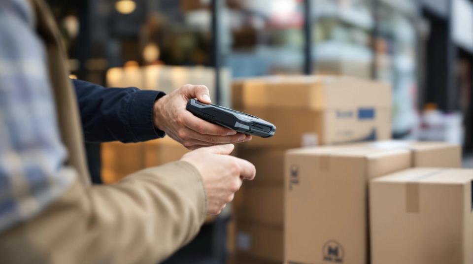 Delivery driver holding out a rugged handheld terminal for a shopkeeper to sign at the doorstep
