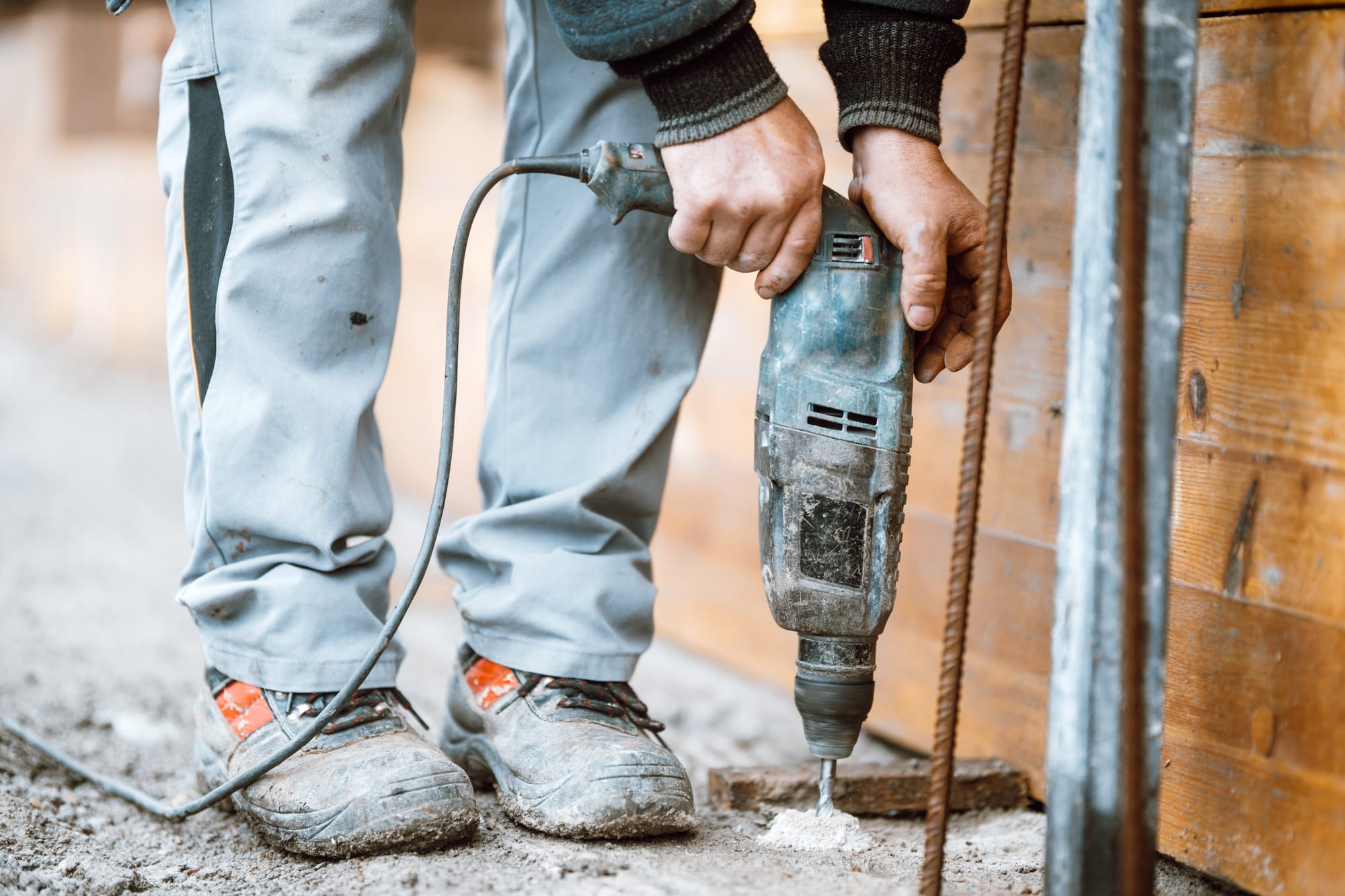 Worker in boots and jeans boring a hole into a grey concrete floor with a rotary hammer, a cone of white drill dust beside the bit