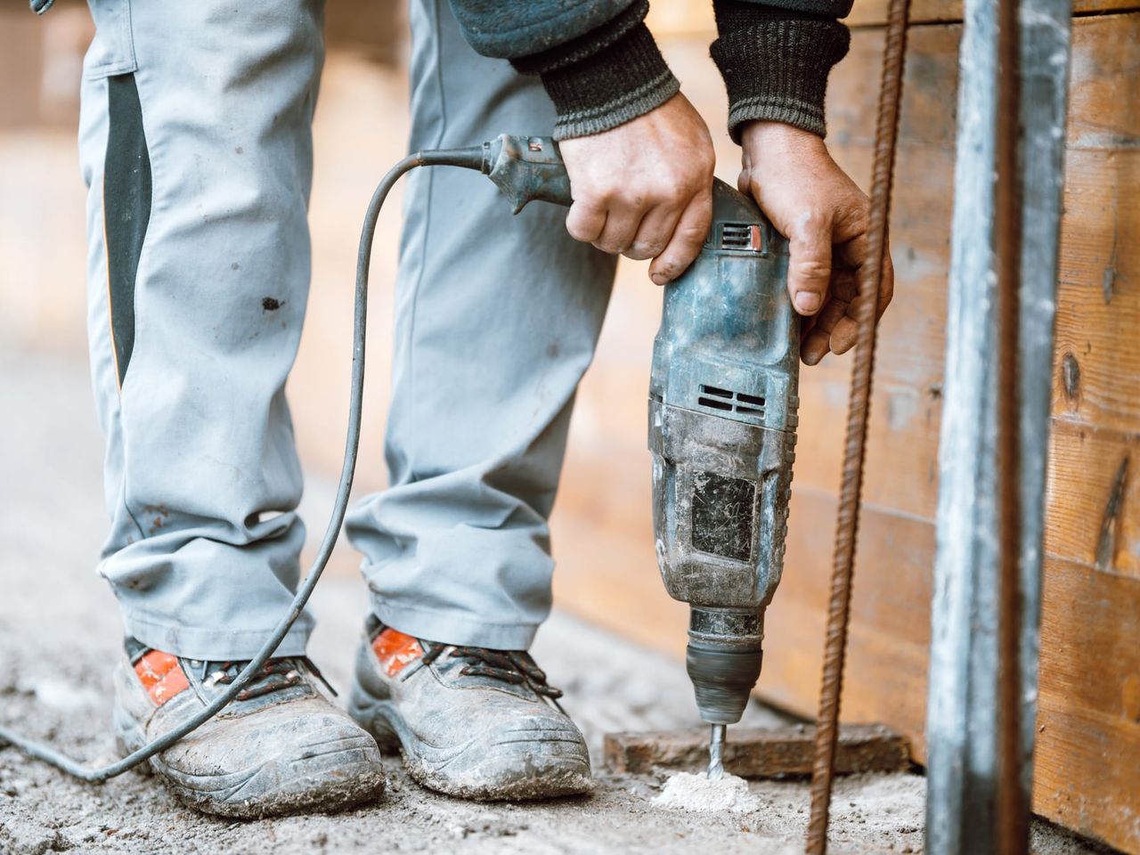 Worker in boots and jeans boring a hole into a grey concrete floor with a rotary hammer, a cone of white drill dust beside the bit