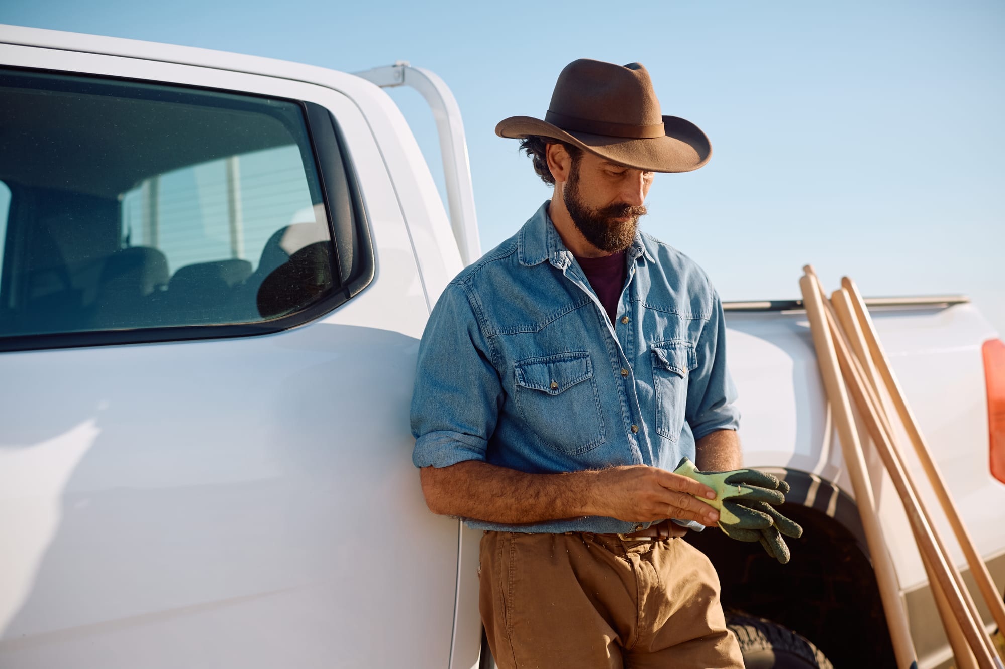 Worker in a denim shirt leaning against the side of a white pickup, pulling on work gloves, stakes stacked in the bed
