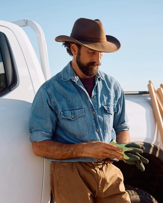 Worker in a denim shirt leaning against the side of a white pickup, pulling on work gloves, stakes stacked in the bed