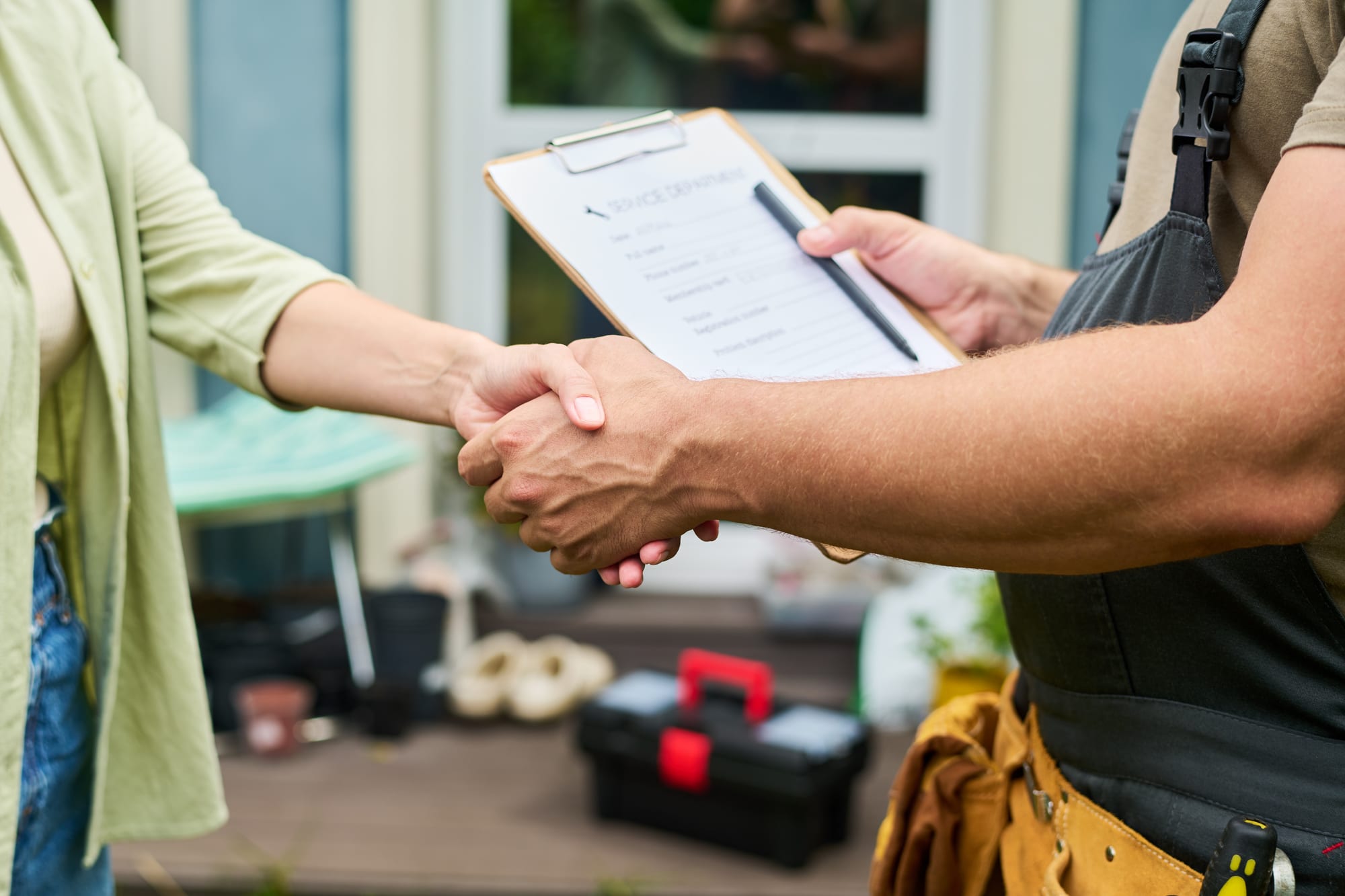 Homeowner shaking hands with a tradesman in bib overalls and a tan leather tool belt, who holds a clipboard and pen, on the deck of a blue-grey house, a toolbox and plant pots behind them