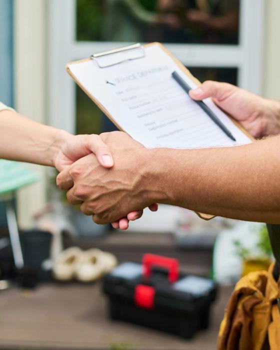 Homeowner shaking hands with a tradesman in bib overalls and a tan leather tool belt, who holds a clipboard and pen, on the deck of a blue-grey house, a toolbox and plant pots behind them