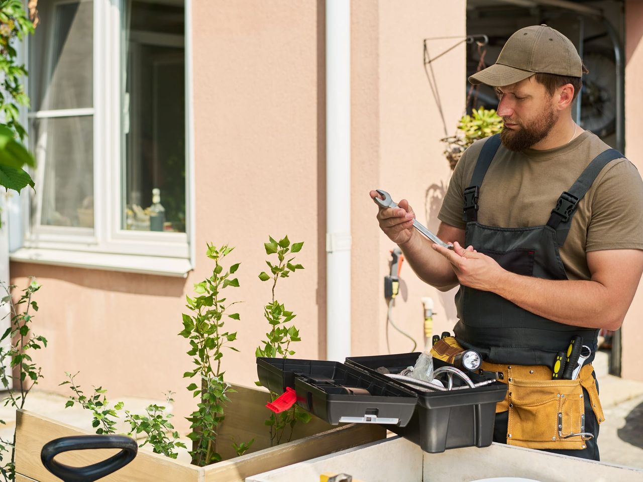 Tradesman in a plain olive T-shirt, bib overalls and a leather tool belt standing at the right of the frame beside an open toolbox outside a house, checking a wrench