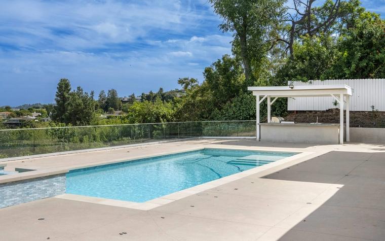 Rectangular backyard swimming pool with a wide poured concrete deck running across the foreground, a board fence and a covered outdoor kitchen behind it