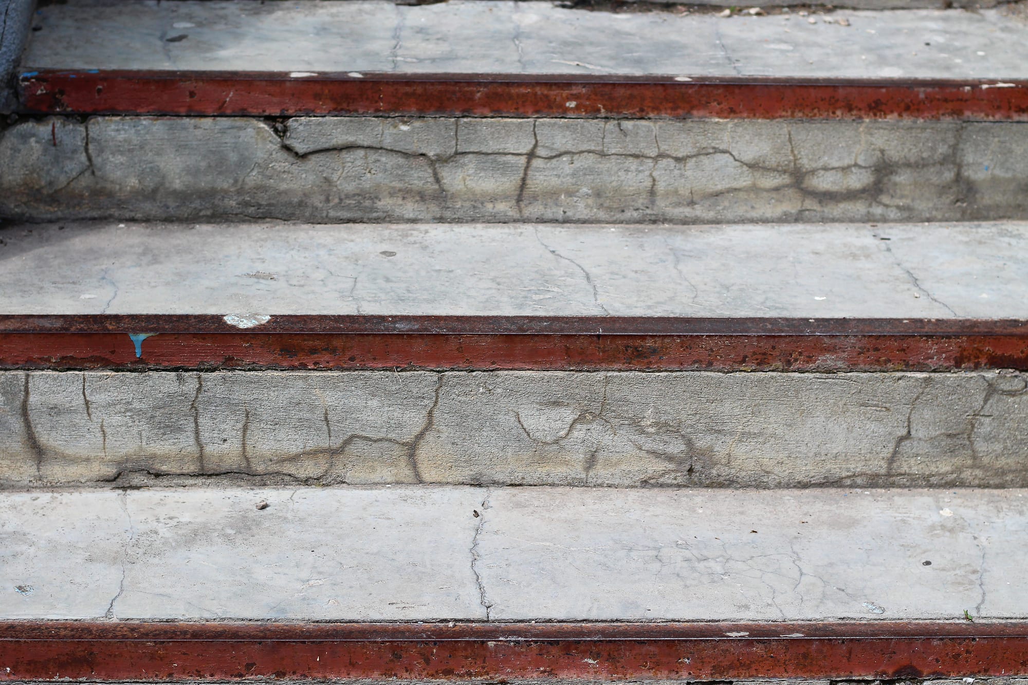Close-up of exterior concrete steps whose treads and risers are broken by a network of cracks, with rusting painted metal nosing strips along each tread