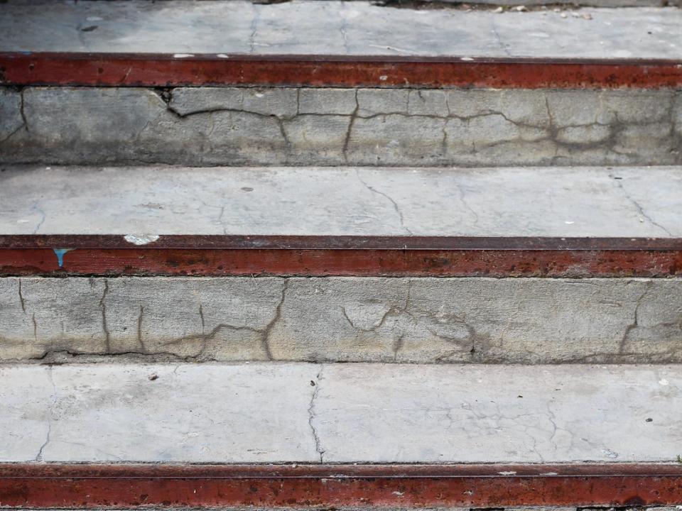 Close-up of exterior concrete steps whose treads and risers are broken by a network of cracks, with rusting painted metal nosing strips along each tread