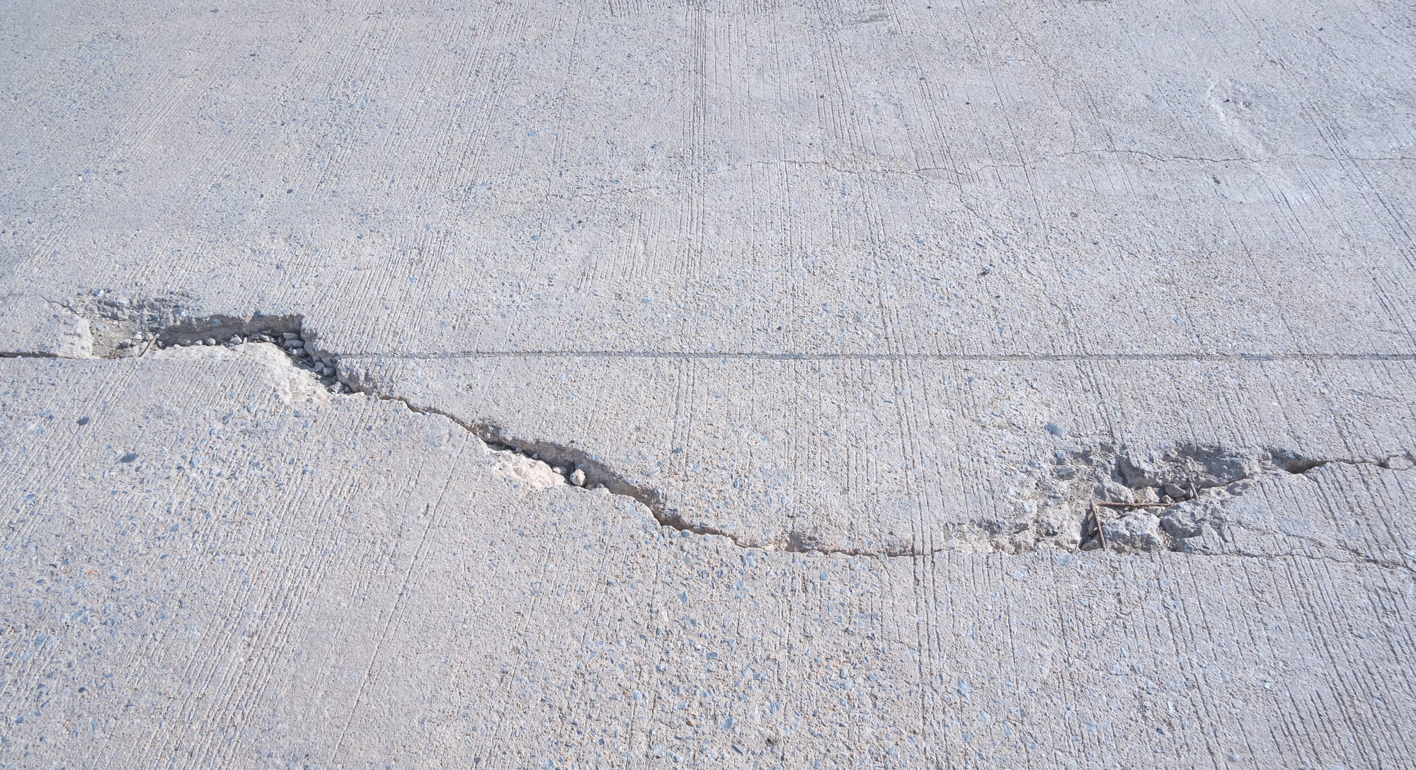 Broom-finished concrete slab where a cracked panel has dropped, leaving a raised lip along the break