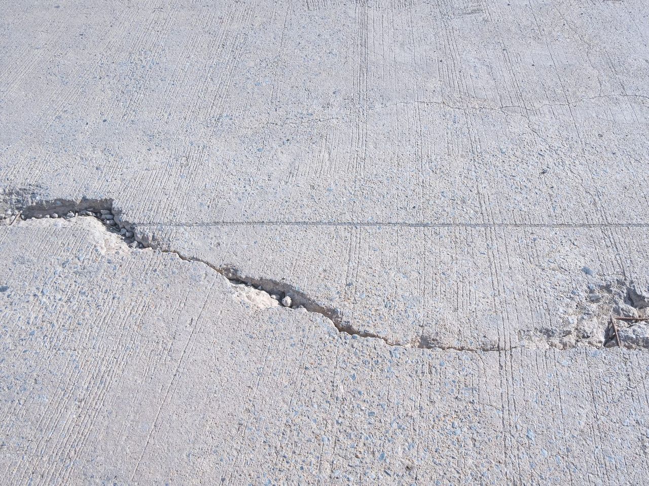 Broom-finished concrete slab where a cracked panel has dropped, leaving a raised lip along the break