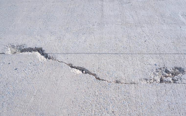 Broom-finished concrete slab where a cracked panel has dropped, leaving a raised lip along the break