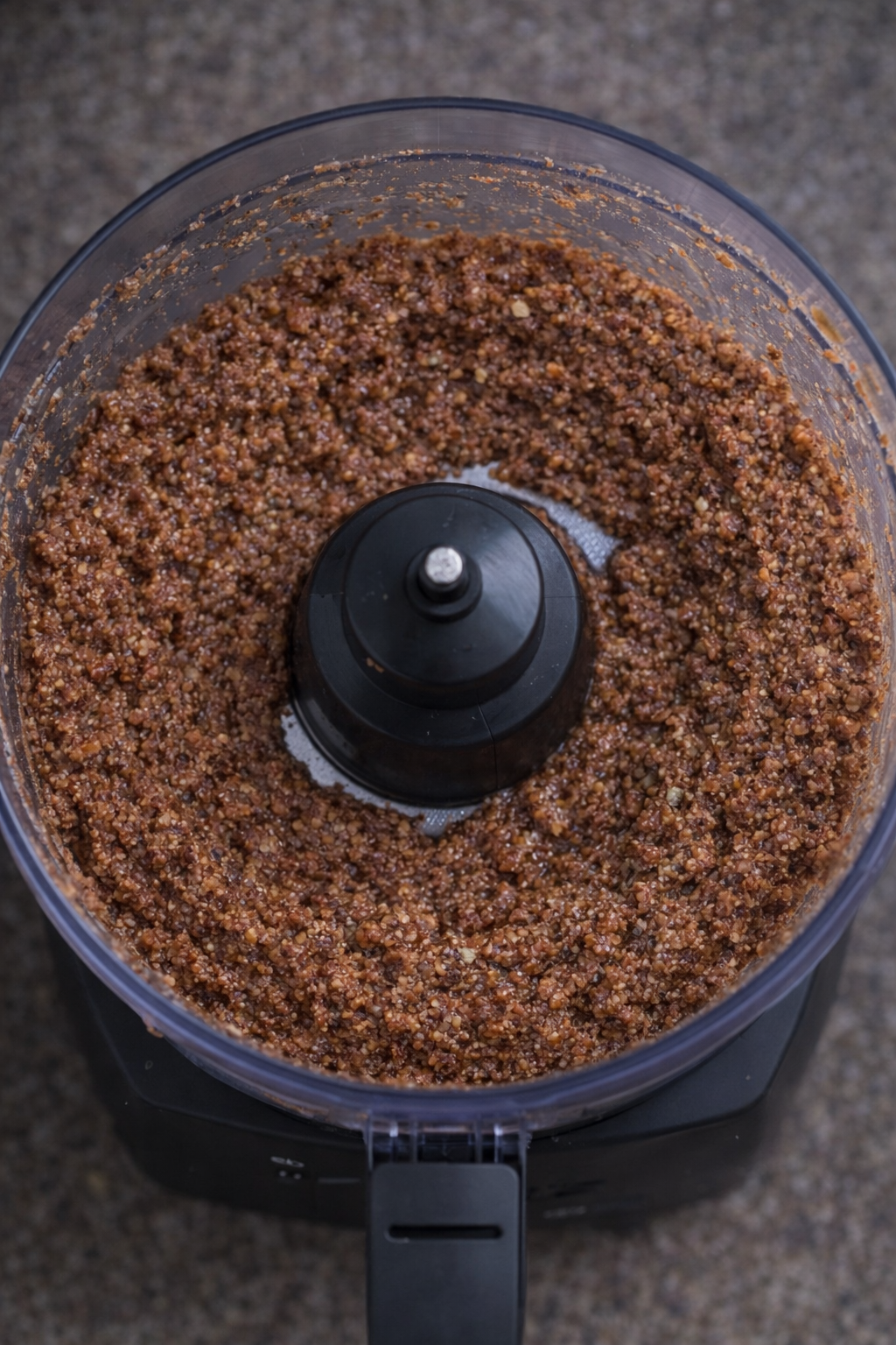 Food processor filled with blended nut and date mixture for crunchy vegan breakfast bars, with spatula scraping down the sides after one minute blitz.