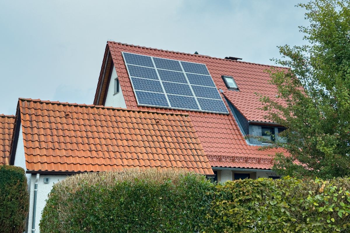 Residential house with red tile roof fitted with rooftop solar panels, partially framed by green hedges and trees under a cloudy sky.