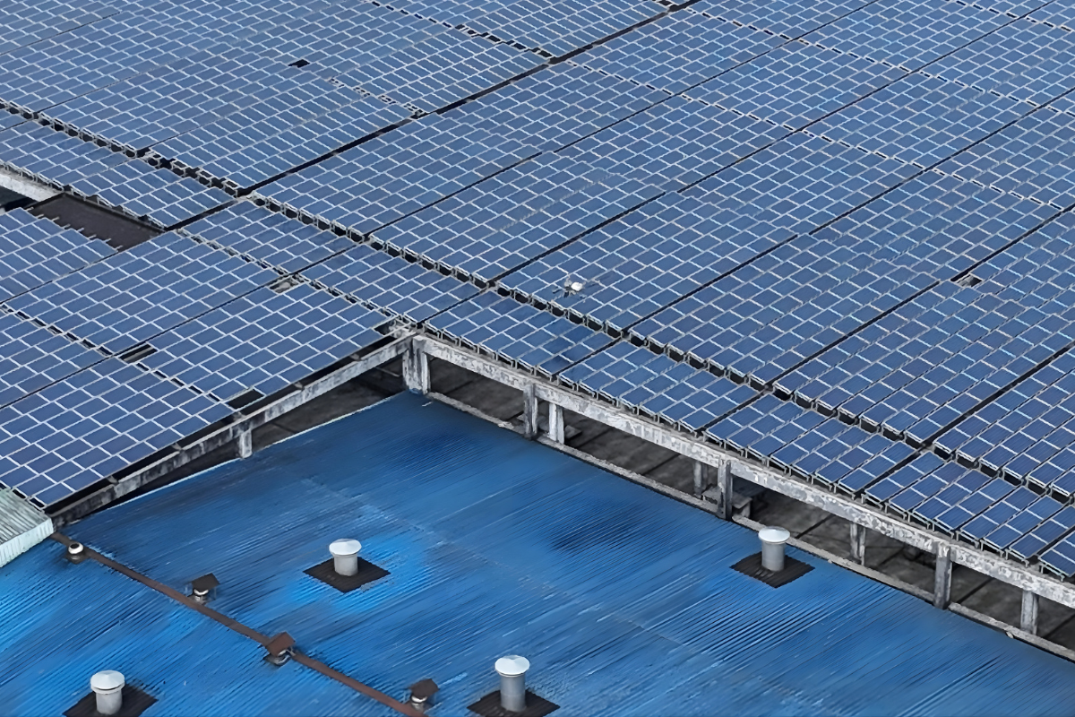 Large array of solar panels installed on the rooftop of a commercial building, with blue metal roofing and ventilation stacks visible below.