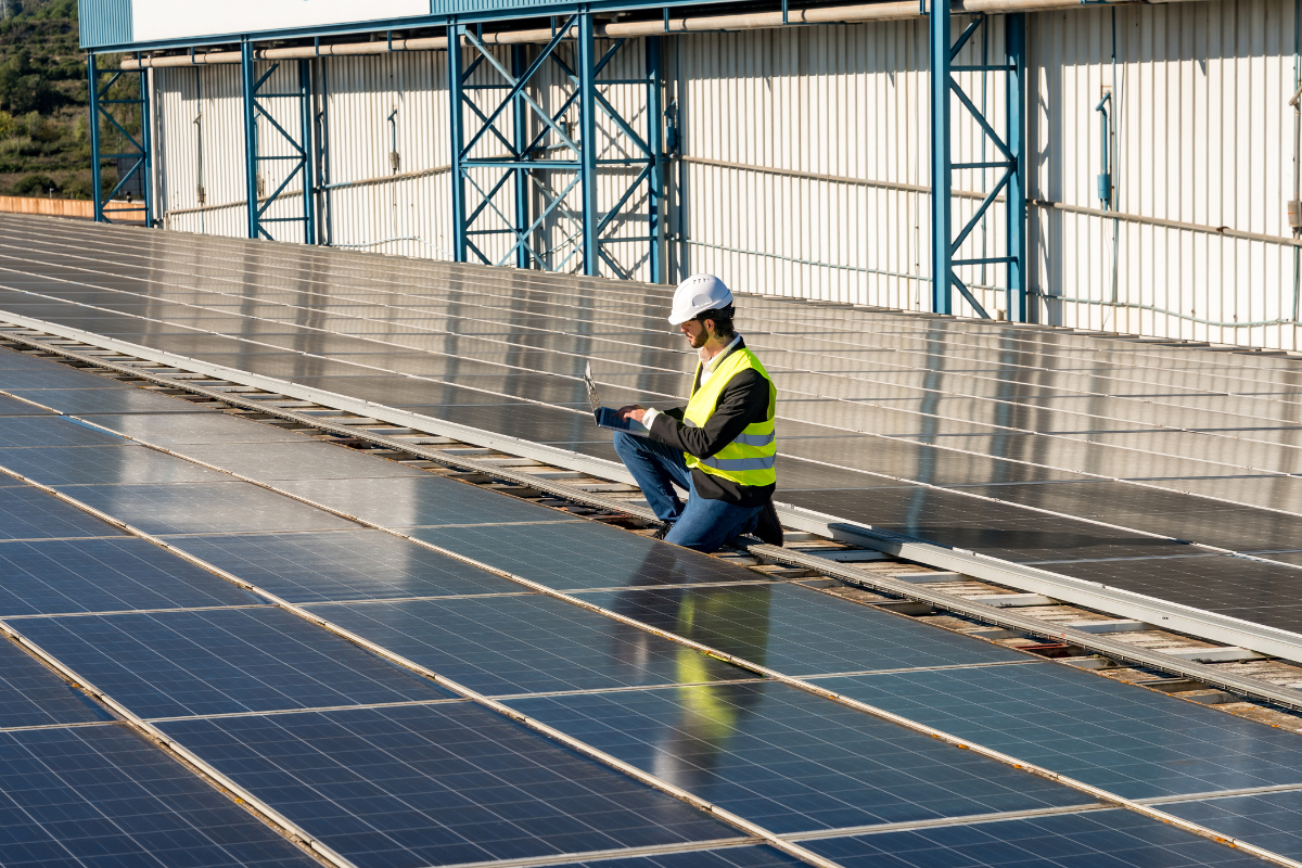 Technician wearing a hard hat and safety vest kneels on a commercial rooftop solar array, using a laptop to inspect rows of solar panels installed on an industrial building.