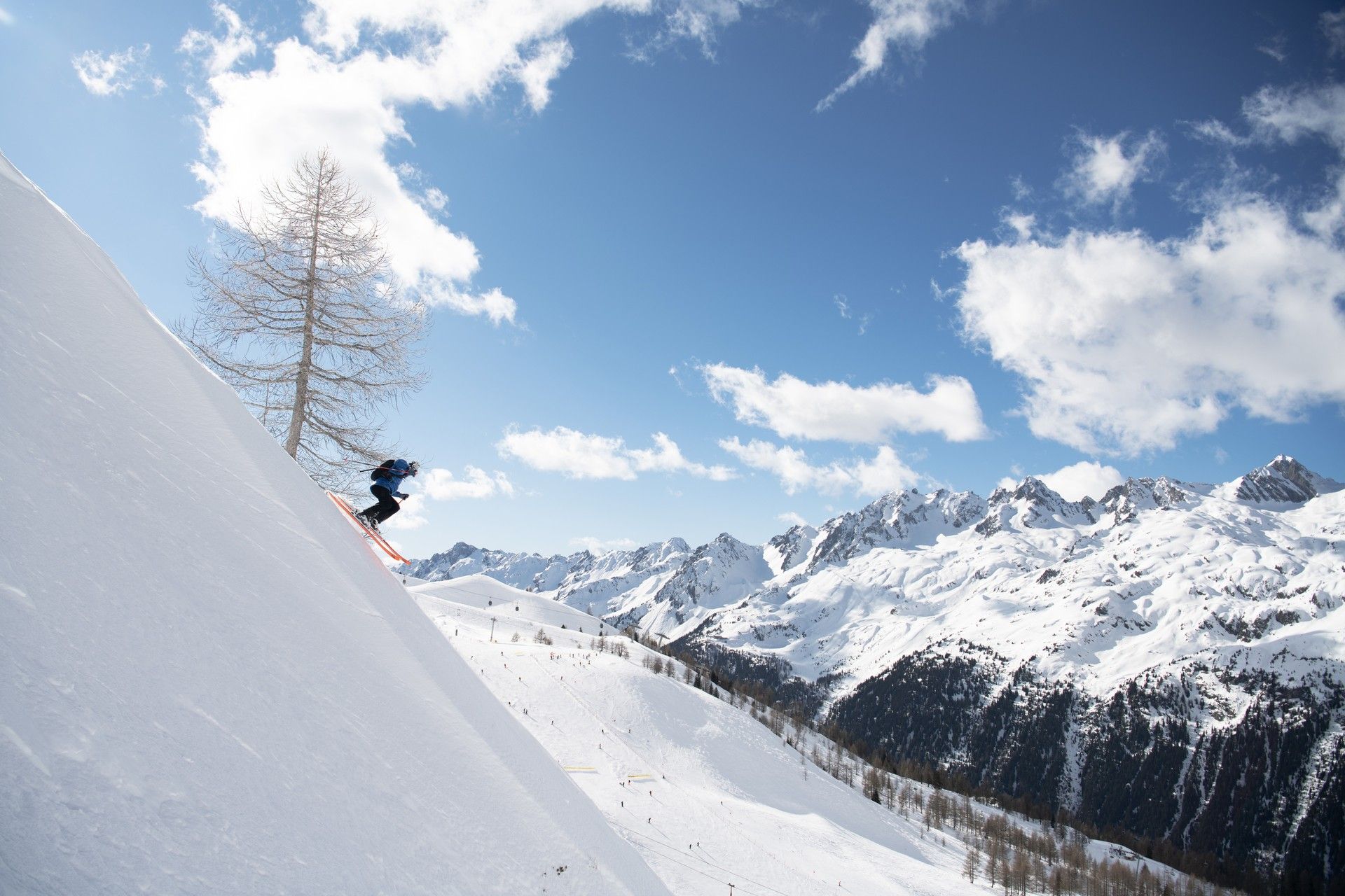 Skier carving down a steep snowy slope with alpine peaks and blue sky in the background