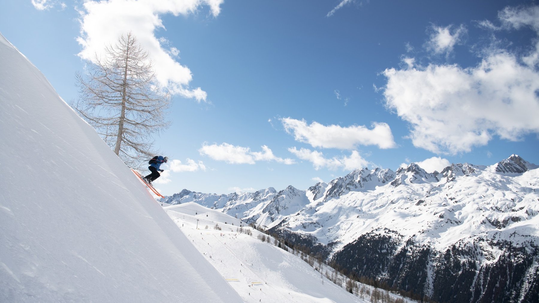 Skier carving down a steep snowy slope with alpine peaks and blue sky in the background