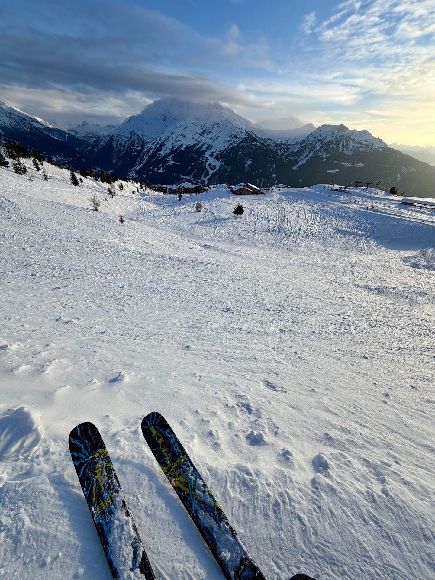 Skier’s view down a snowy alpine slope with ski tracks and mountain peaks under blue sky.