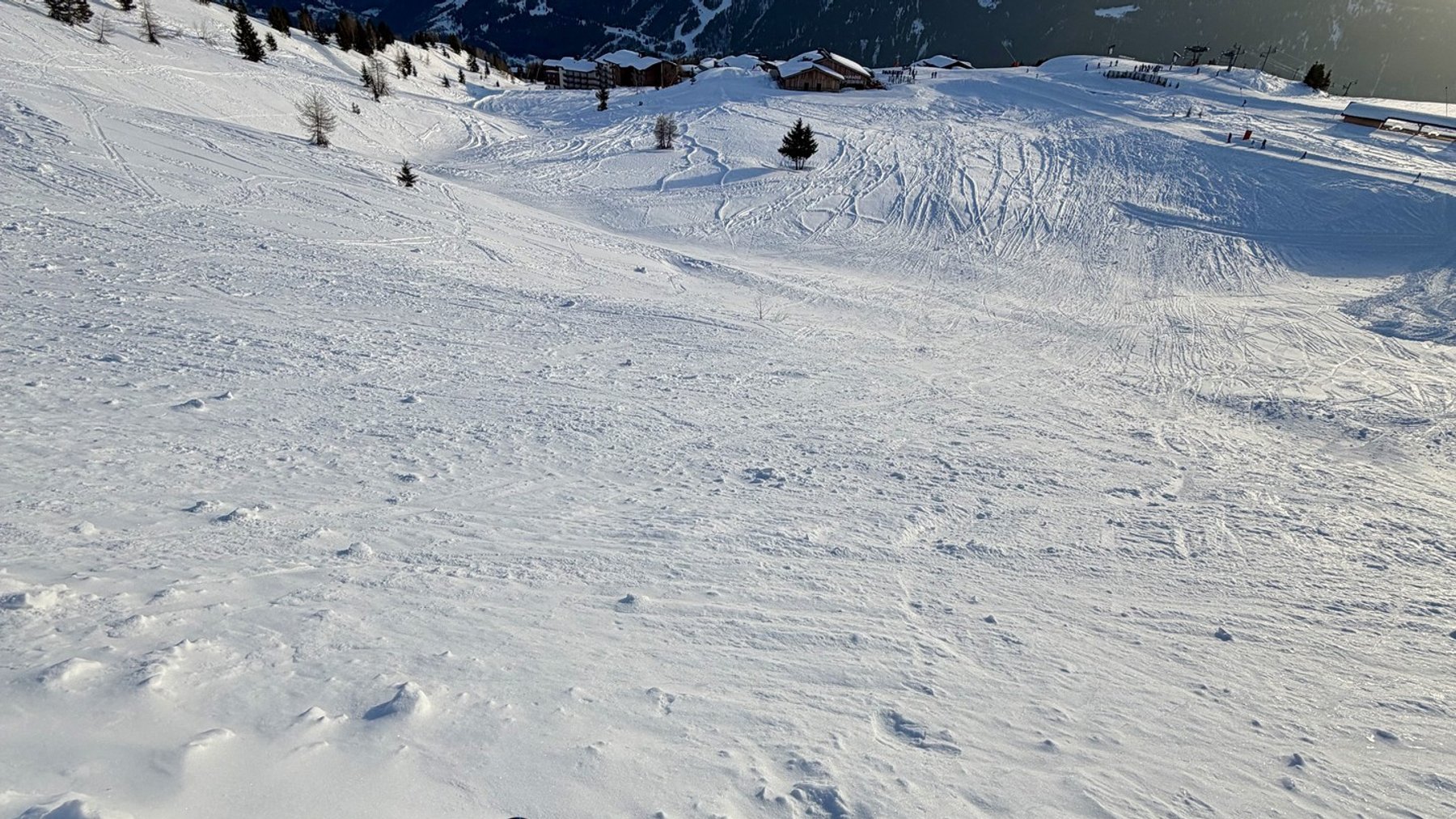 Skier’s view down a snowy alpine slope with ski tracks and mountain peaks under blue sky.