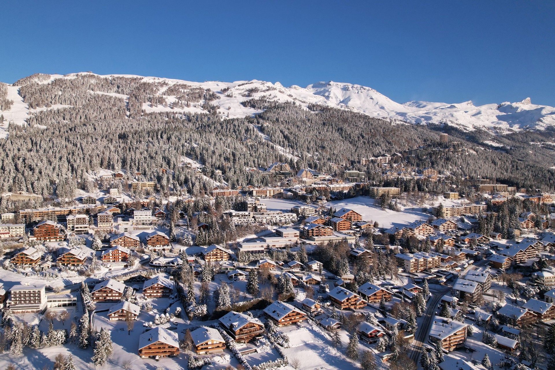 Snow-covered alpine village with mountains and ski slopes under a clear blue sky.