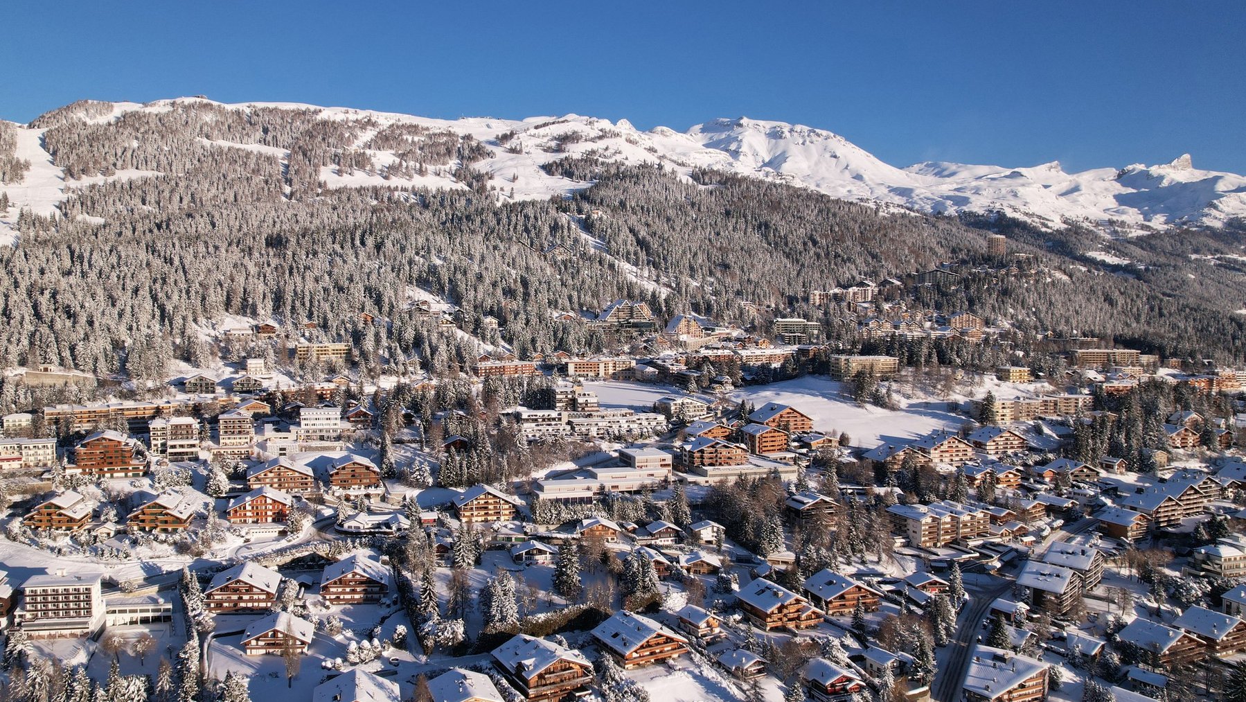 Snow-covered alpine village with mountains and ski slopes under a clear blue sky.