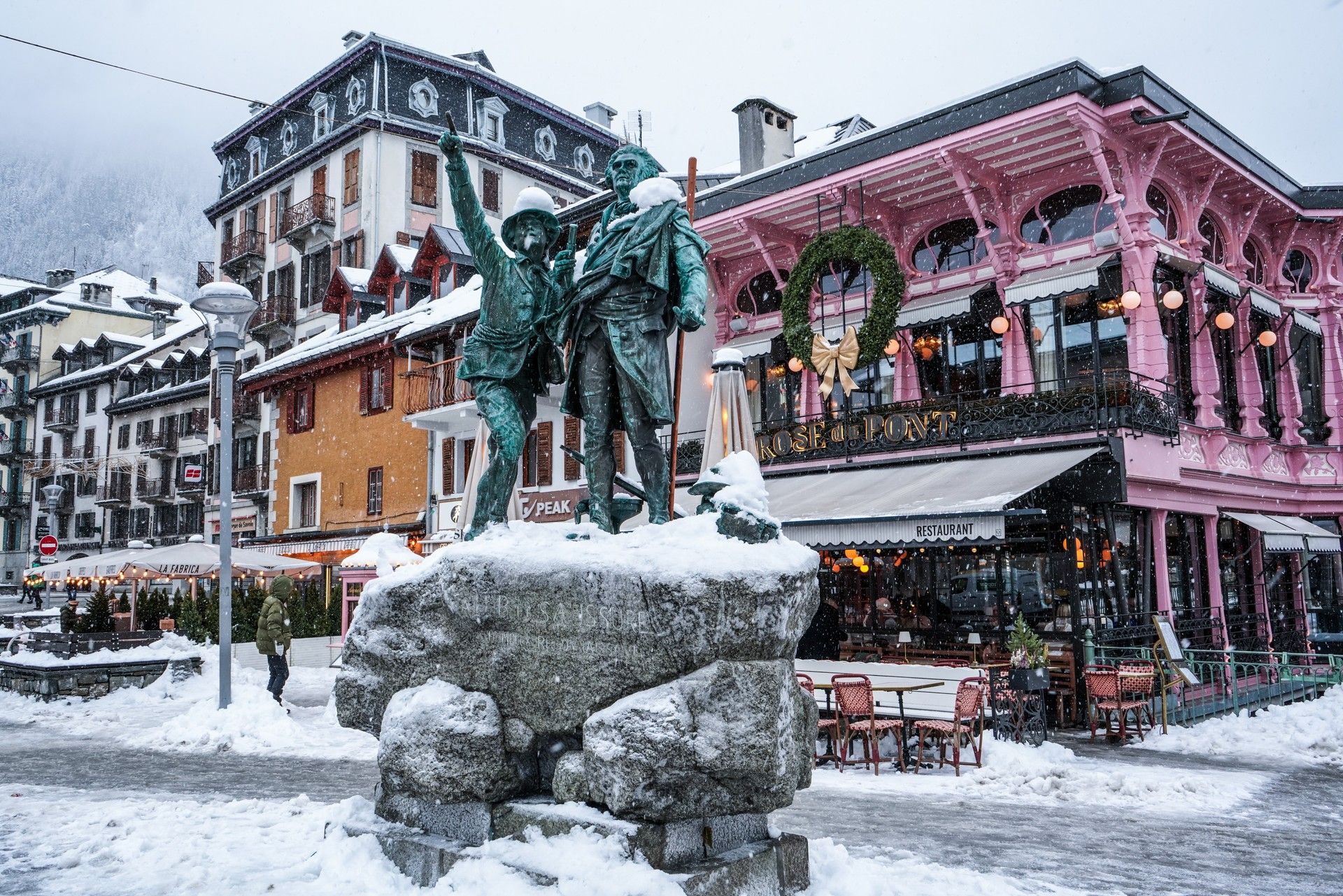 Snowy mountain square with a bronze statue in front of pastel shops and cafes