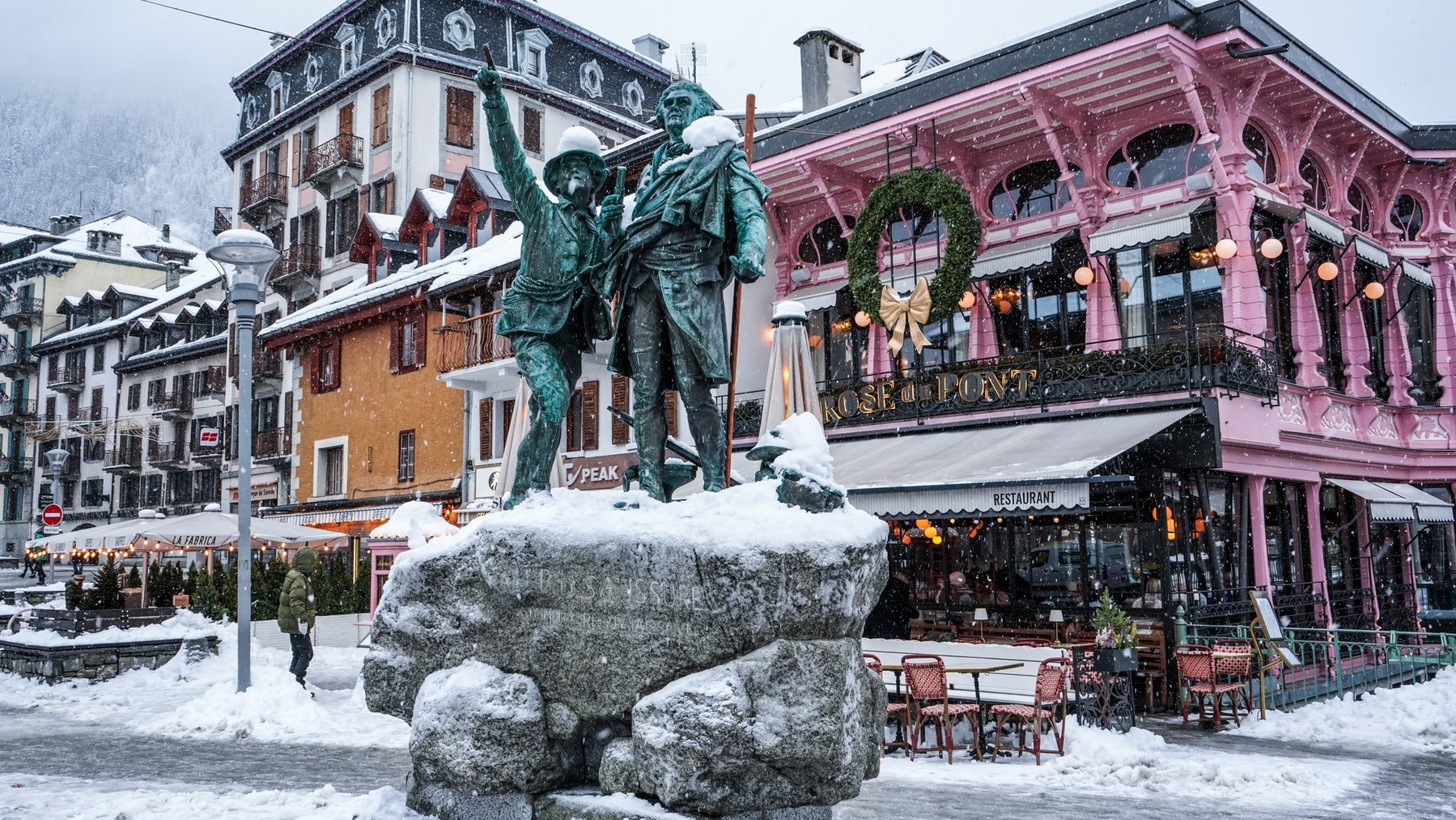 Snowy mountain square with a bronze statue in front of pastel shops and cafes