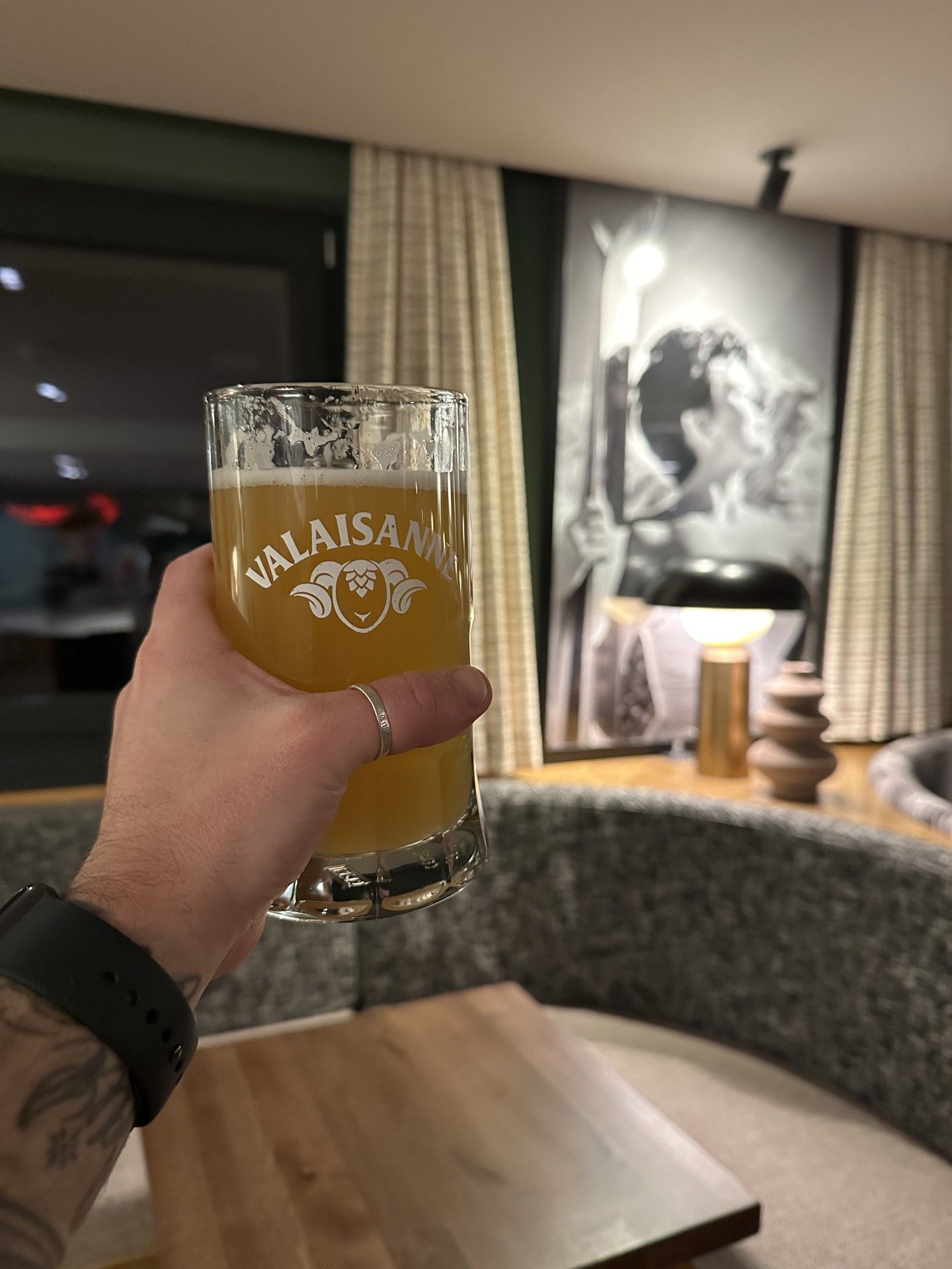 Hand holds a beer glass labeled “VALAISAN” in a cozy lounge bar setting