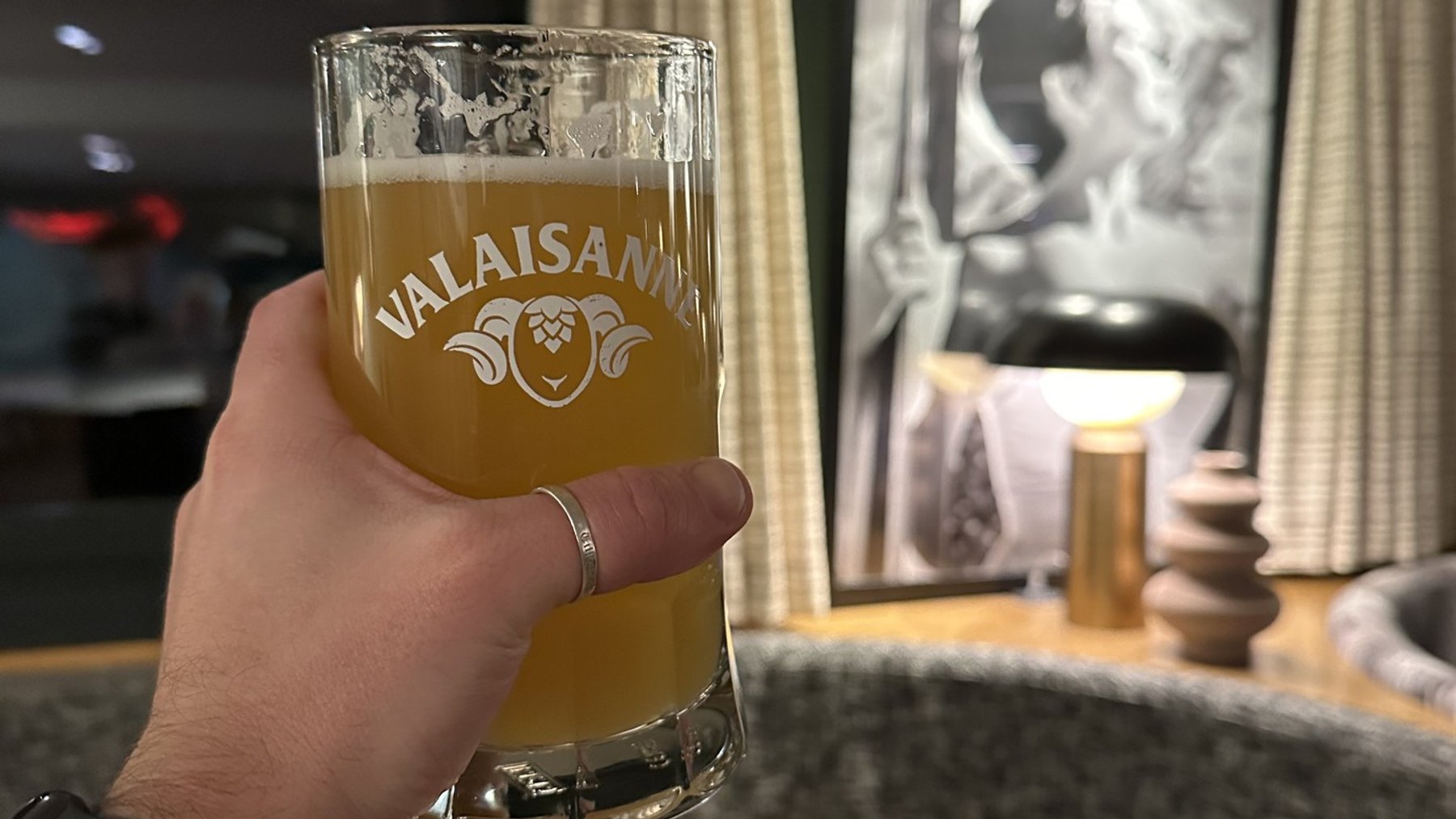 Hand holds a beer glass labeled “VALAISAN” in a cozy lounge bar setting