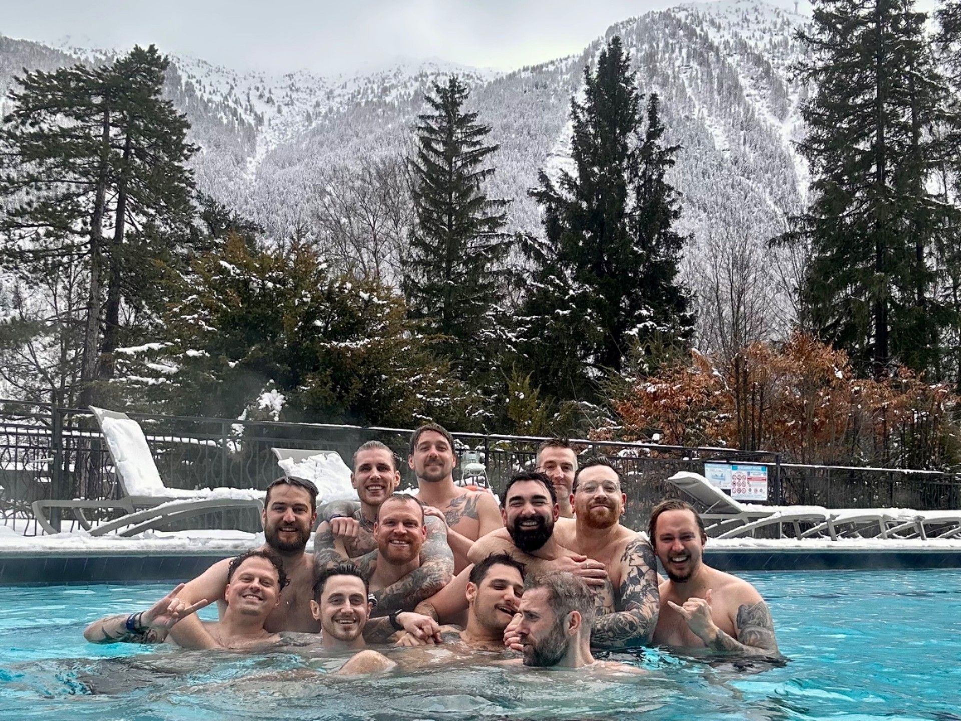 A group of adults pose in an outdoor pool with snow-covered mountains and evergreen trees behind them.
