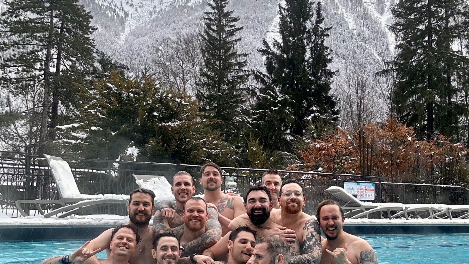 A group of adults pose in an outdoor pool with snow-covered mountains and evergreen trees behind them.