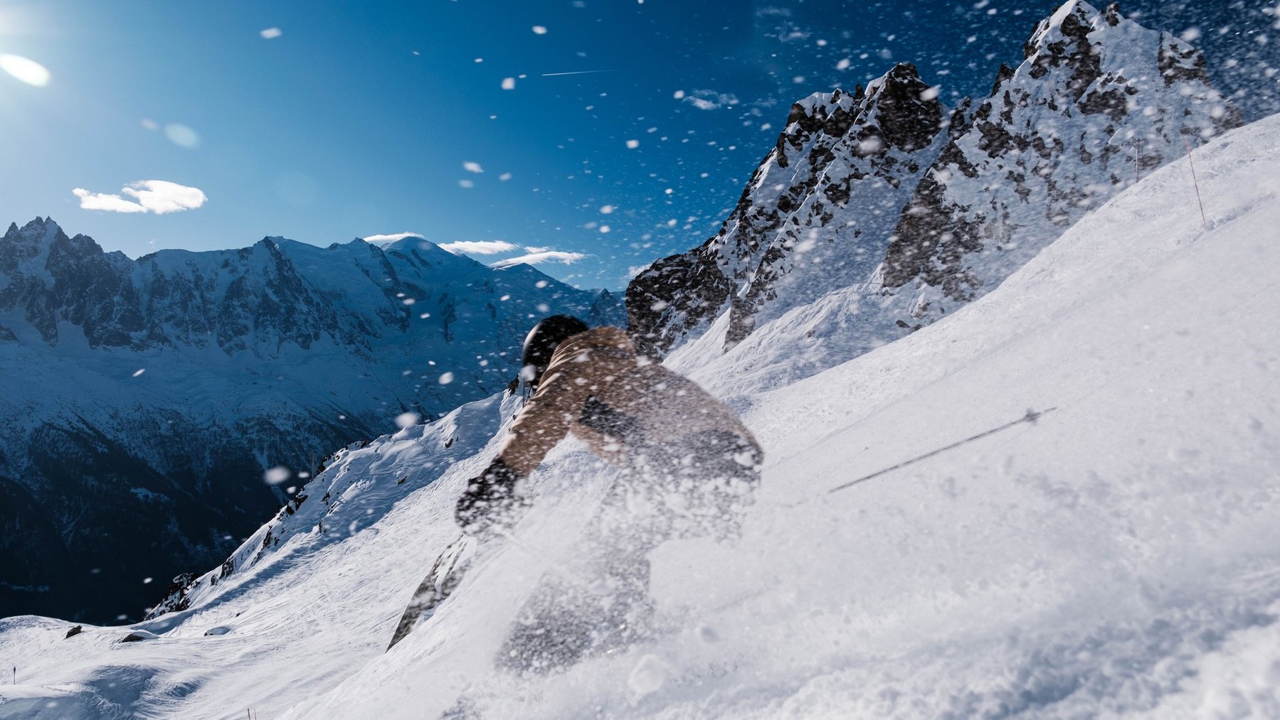 Skiers carve across snowy alpine slope as powder sprays from a dramatic rightward jump.