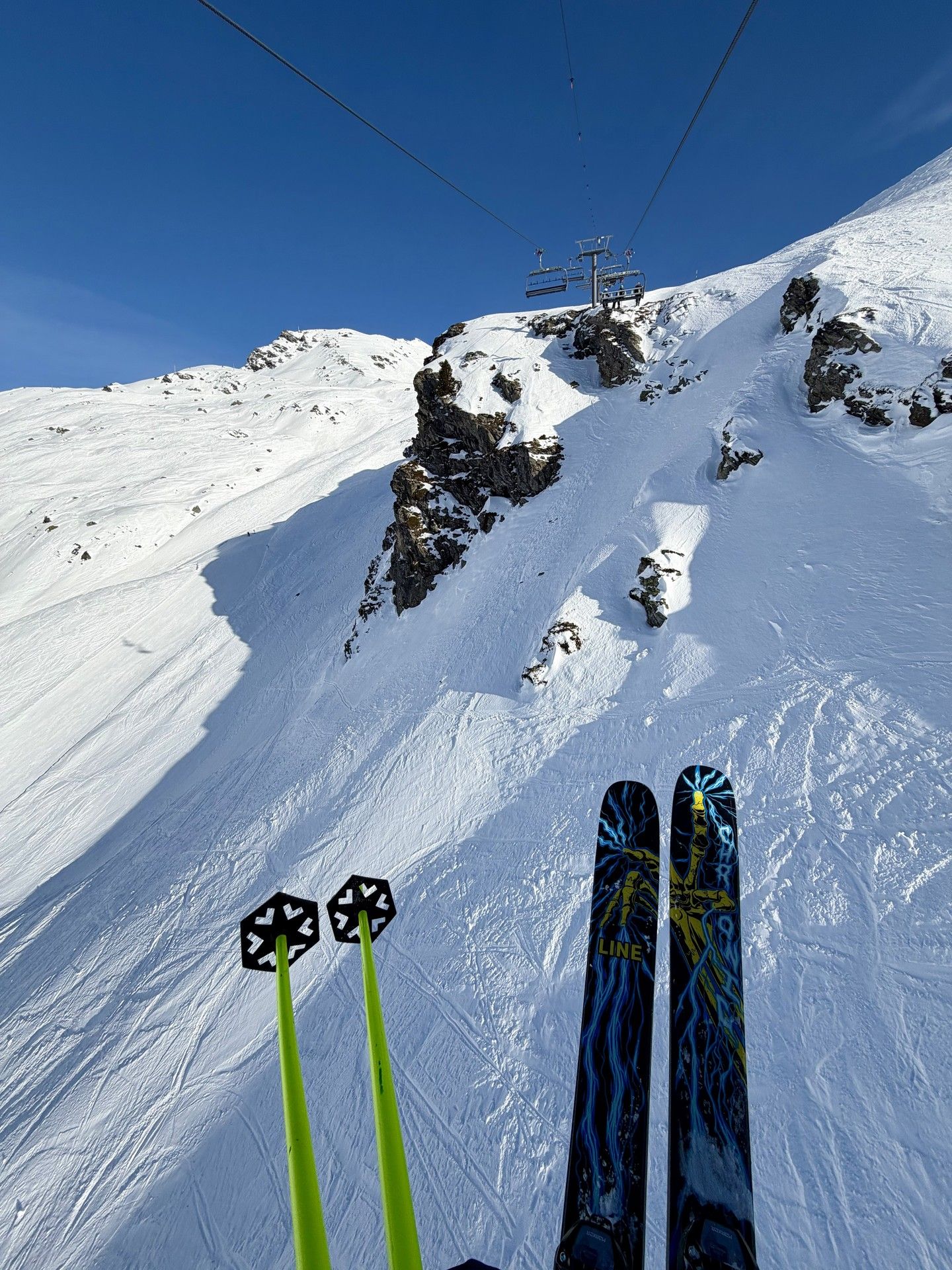 Skiers descend a snowy mountain slope toward ski lift equipment under bright blue skies.