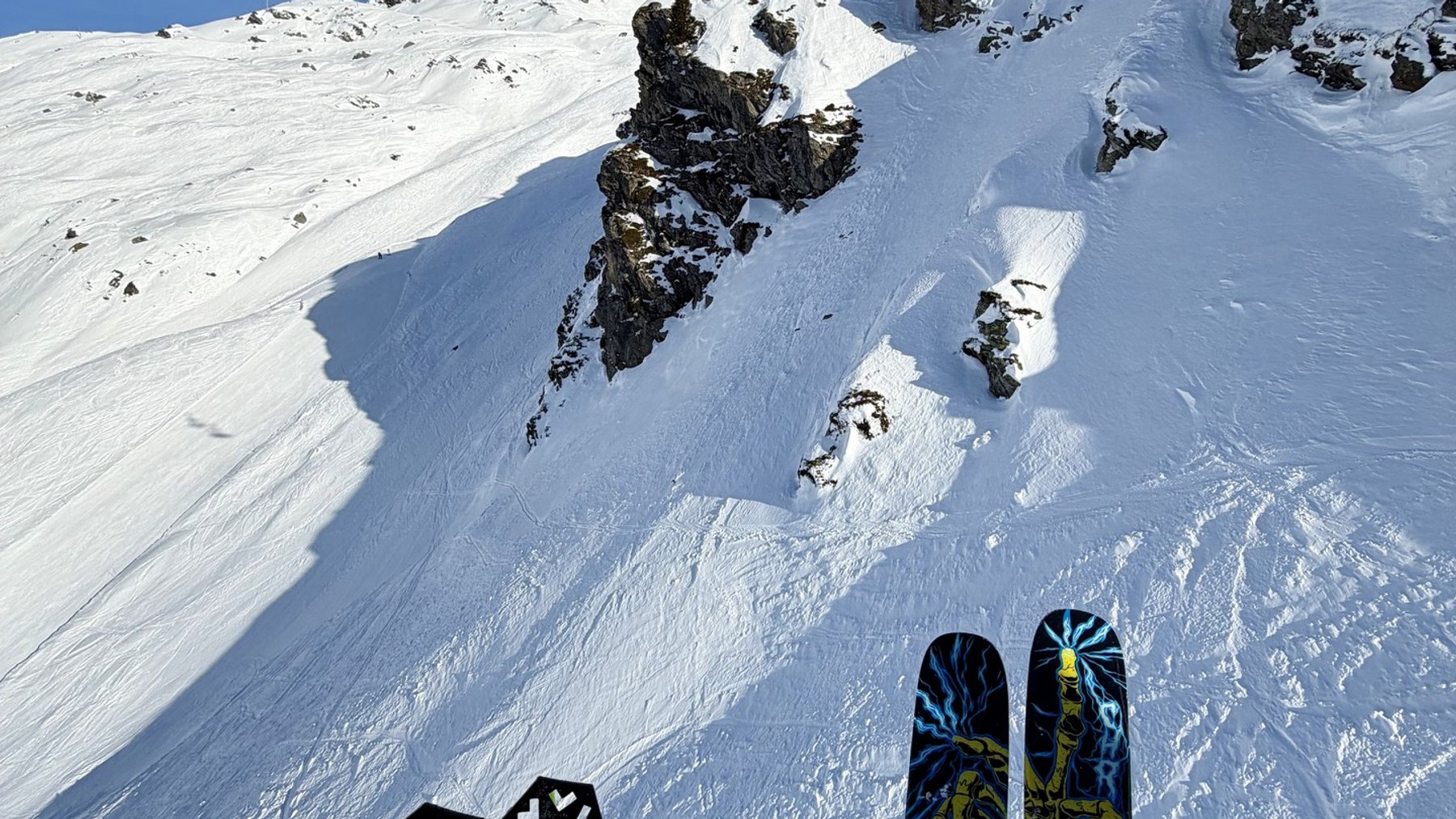 Skiers descend a snowy mountain slope toward ski lift equipment under bright blue skies.