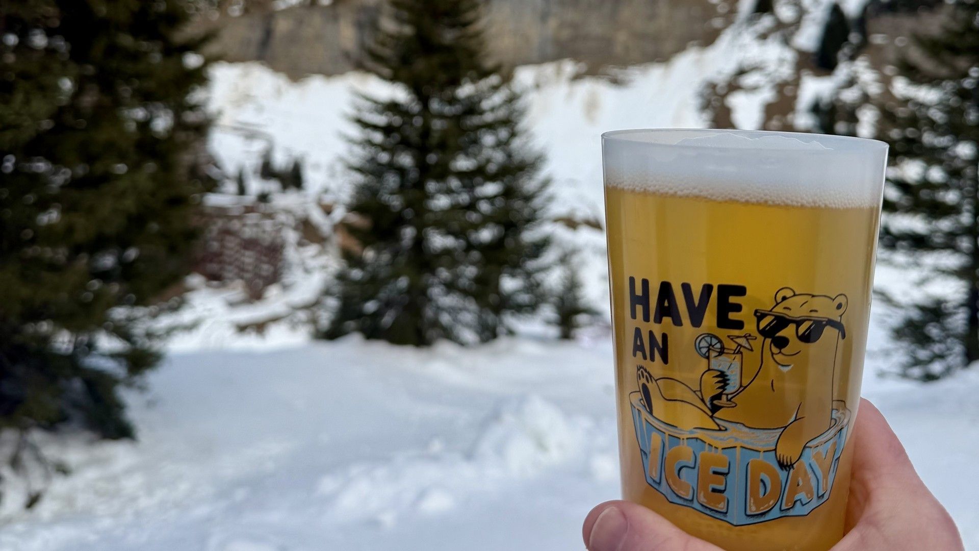 Hand holds a yellow “Have an Ice Day” drink cup in snowy alpine landscape