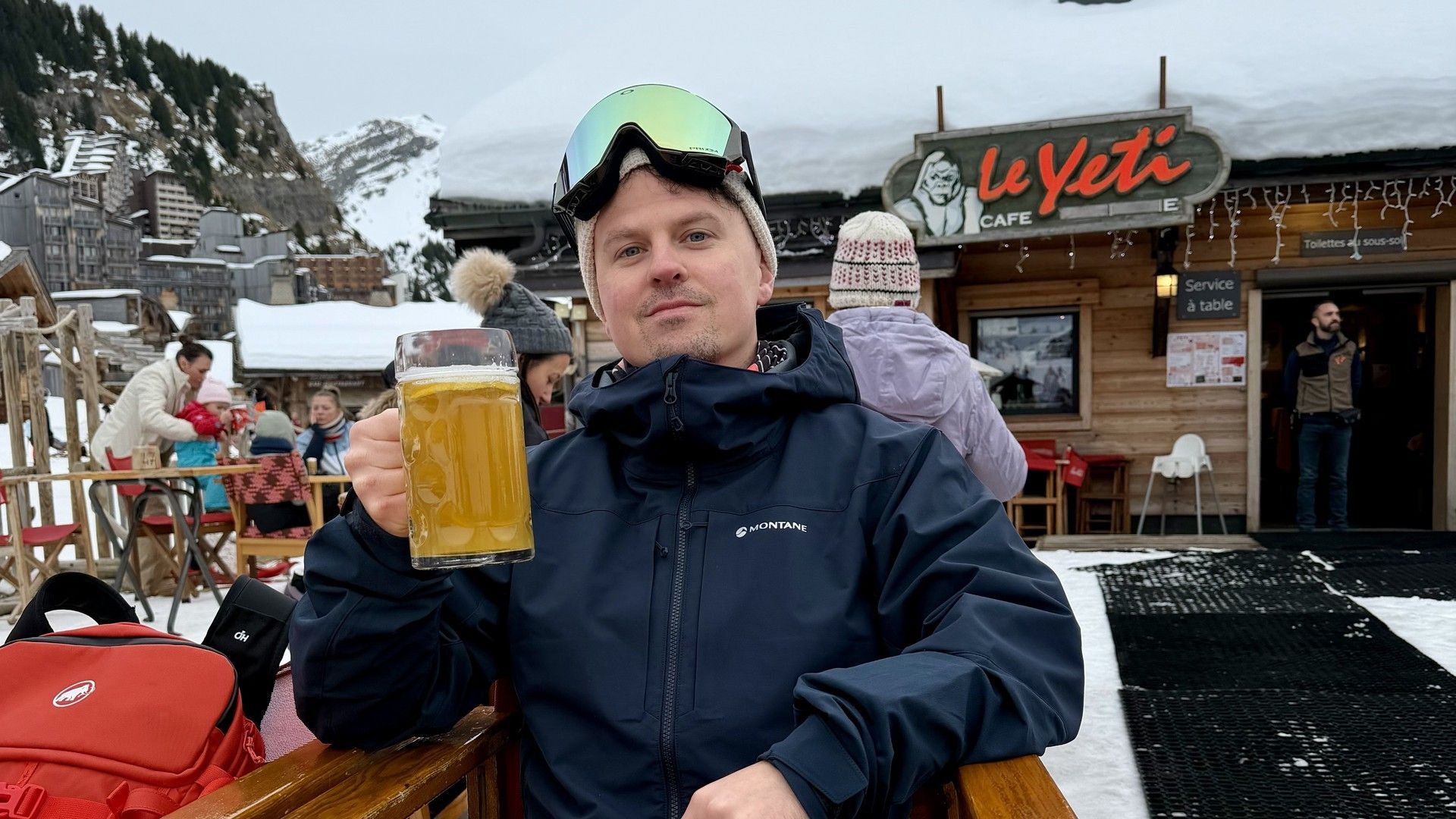 Man in ski jacket and sunglasses holding a mug at a snowy mountain restaurant patio
