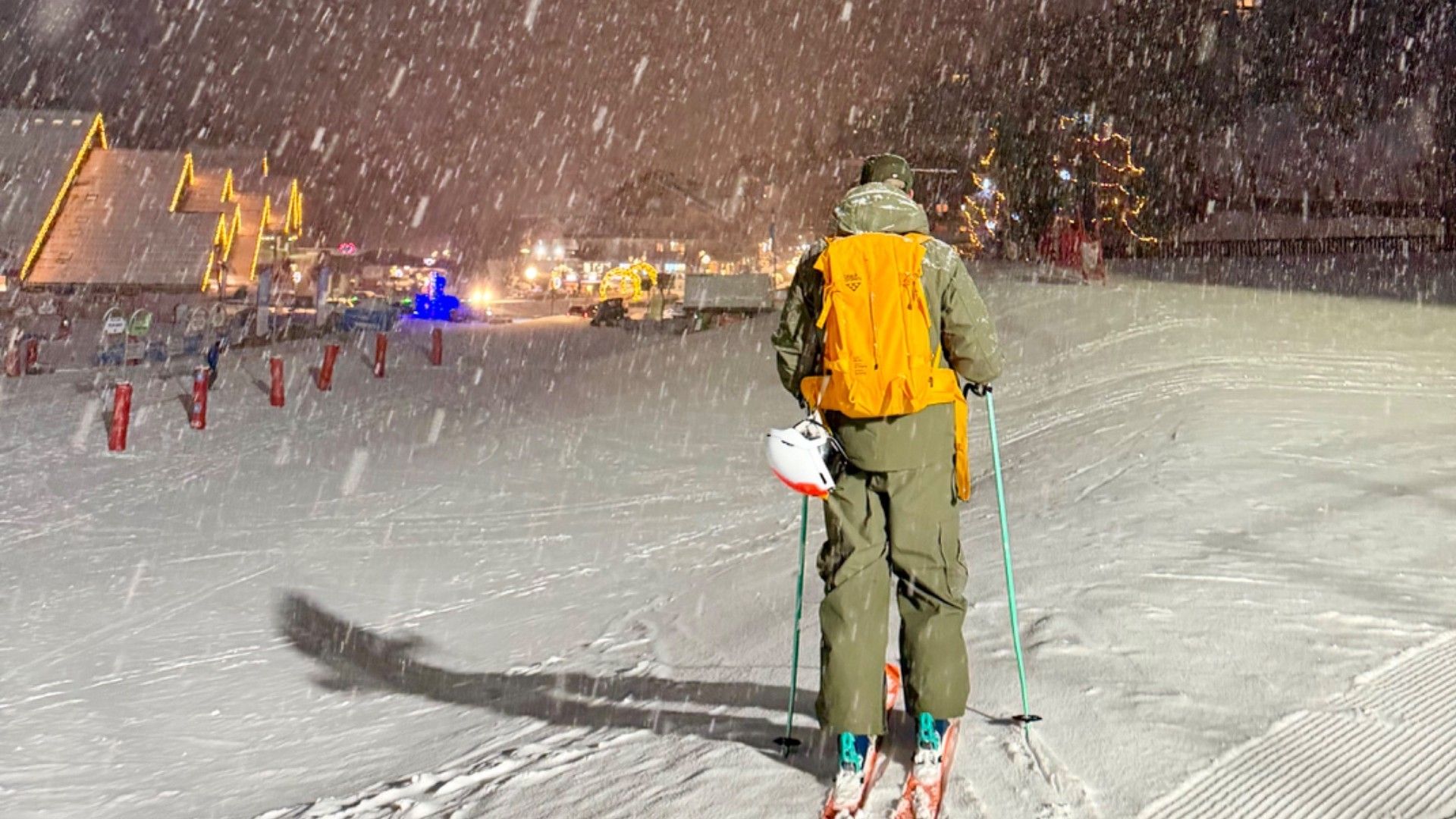 A skier descends a snowy piste at night as heavy snow falls and resort lights glow in the distance.