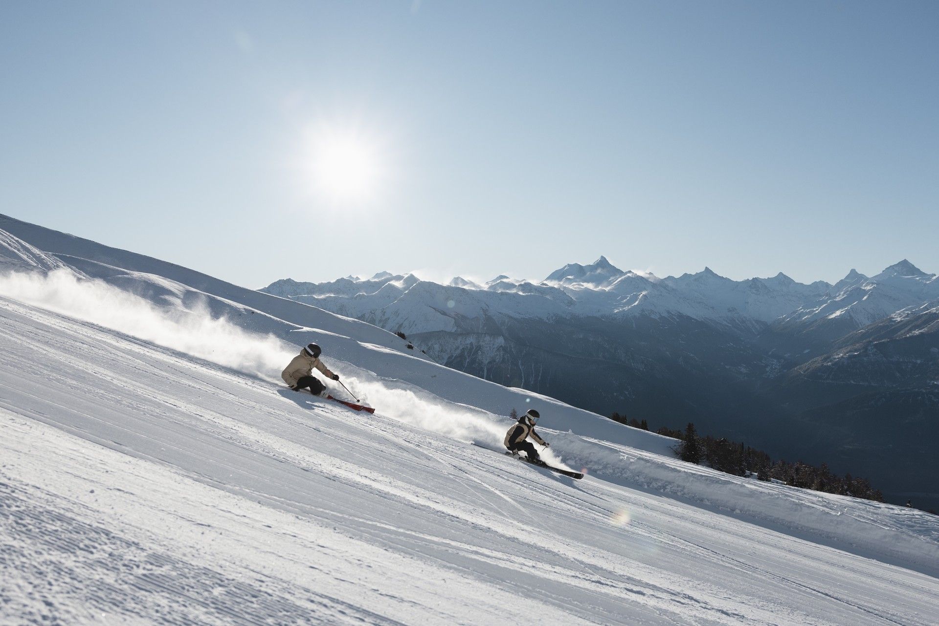 Two skiers carve down a snowy slope toward mountain peaks under bright sunshine