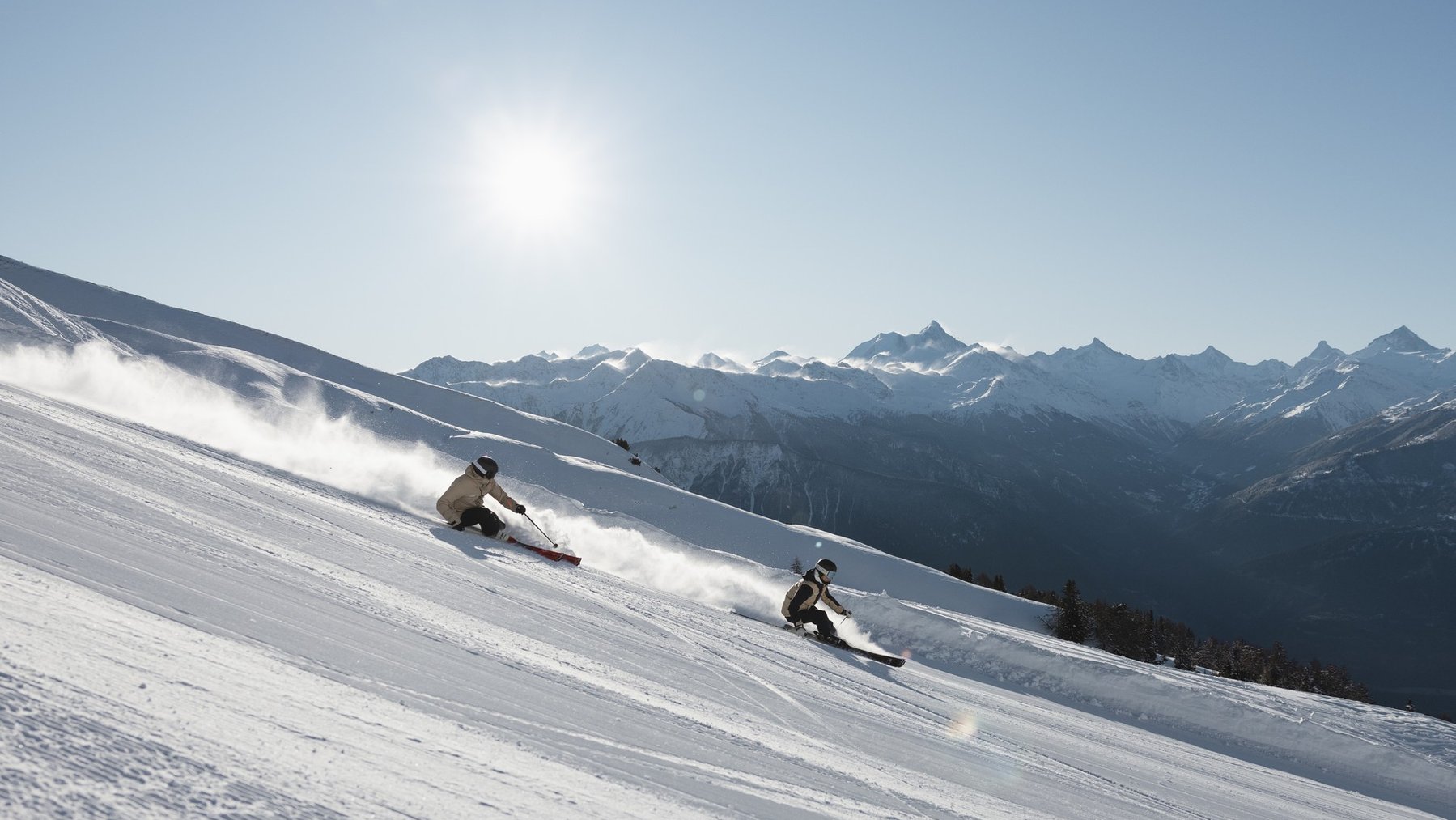 Two skiers carve down a snowy slope toward mountain peaks under bright sunshine