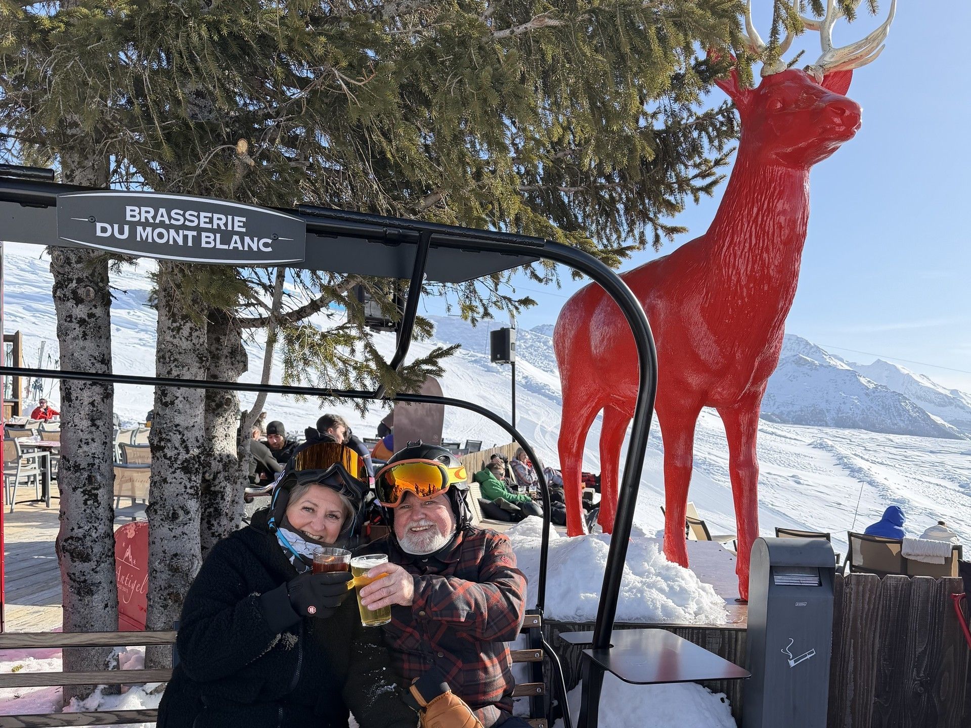 Two skiers sitting on a bench with drinks in front of a red reindeer sculpture near a ski station sign