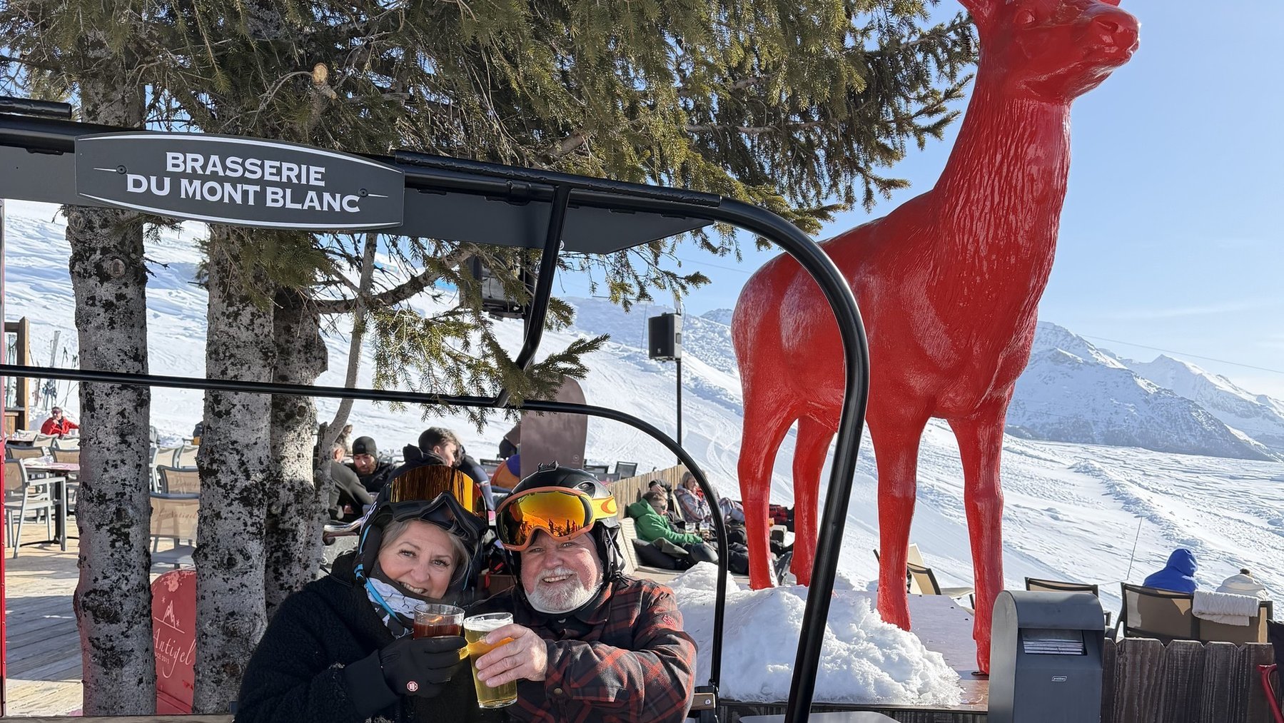 Two skiers sitting on a bench with drinks in front of a red reindeer sculpture near a ski station sign