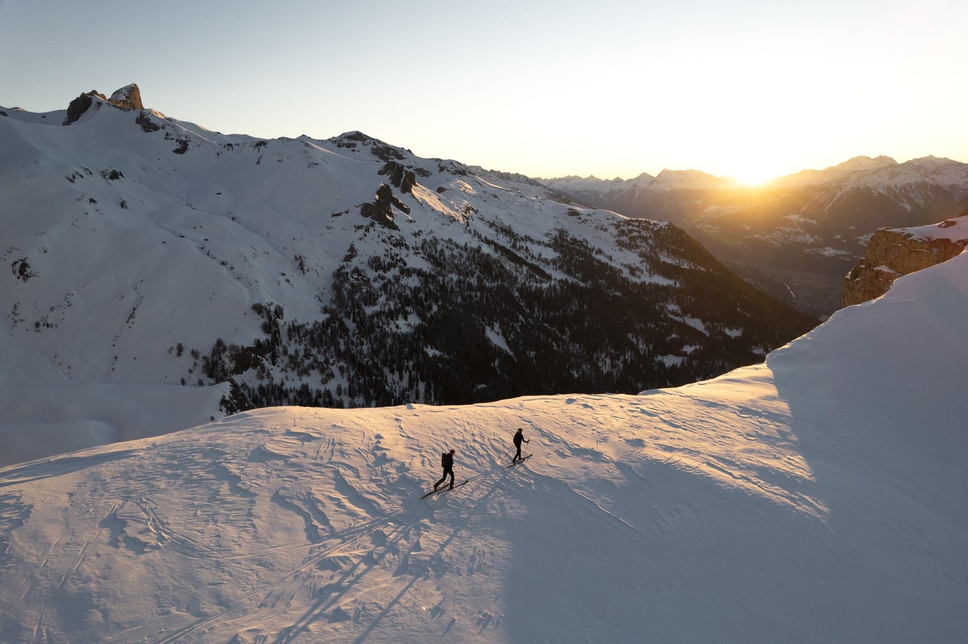 Two skiers trek across a snowy alpine slope at sunrise between mountain peaks
