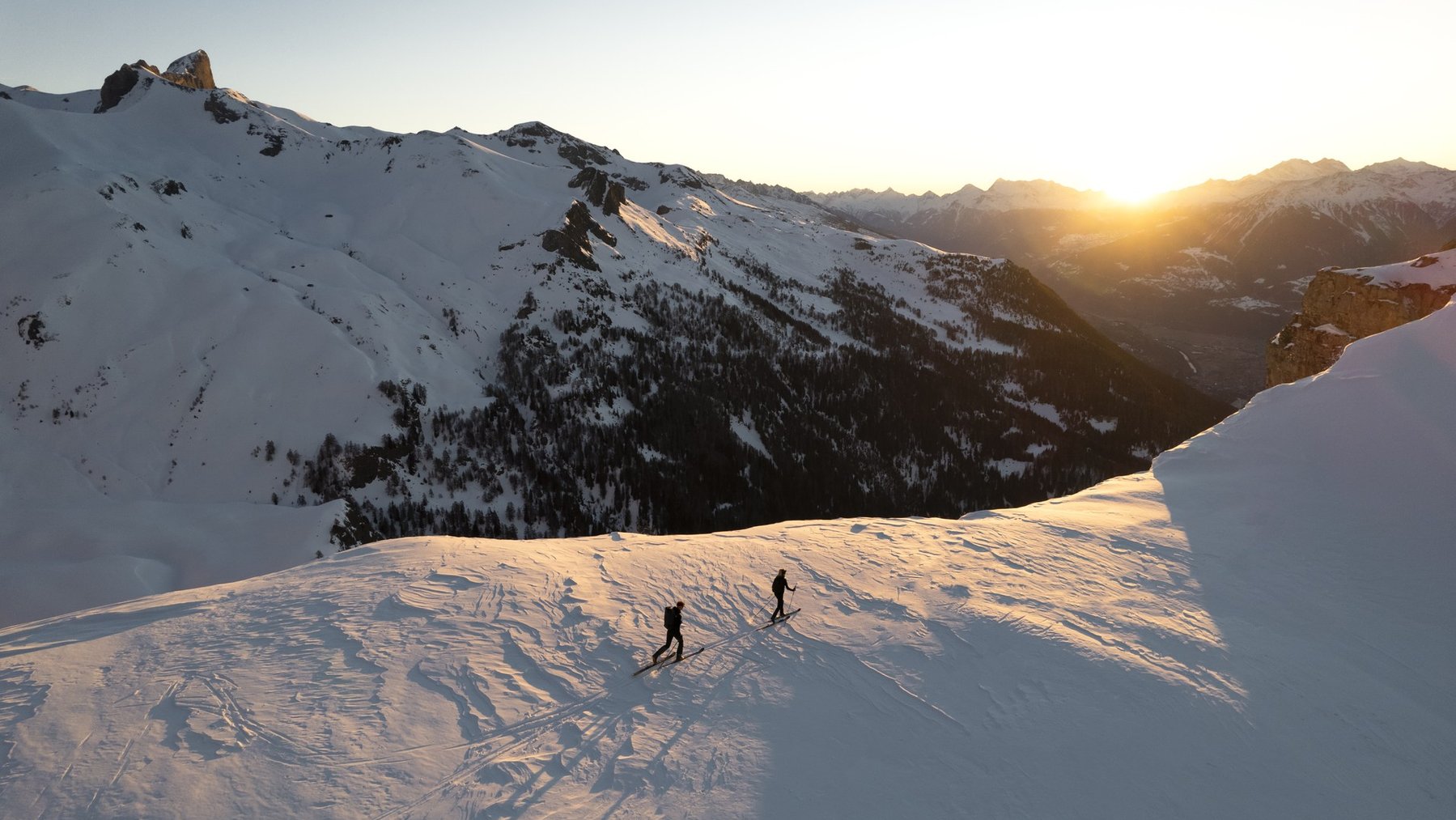 Two skiers trek across a snowy alpine slope at sunrise between mountain peaks