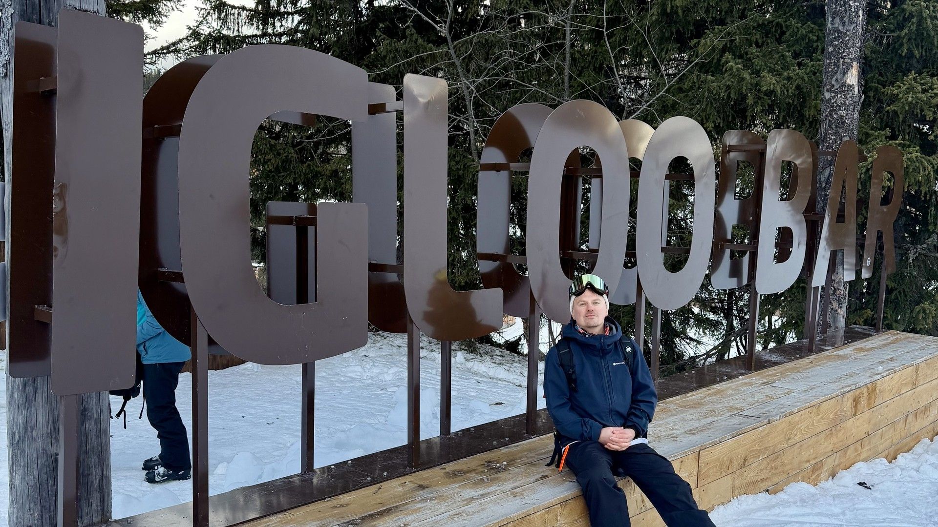 Man sitting on snowy steps in front of giant “GLOBIX” letters at a winter resort.