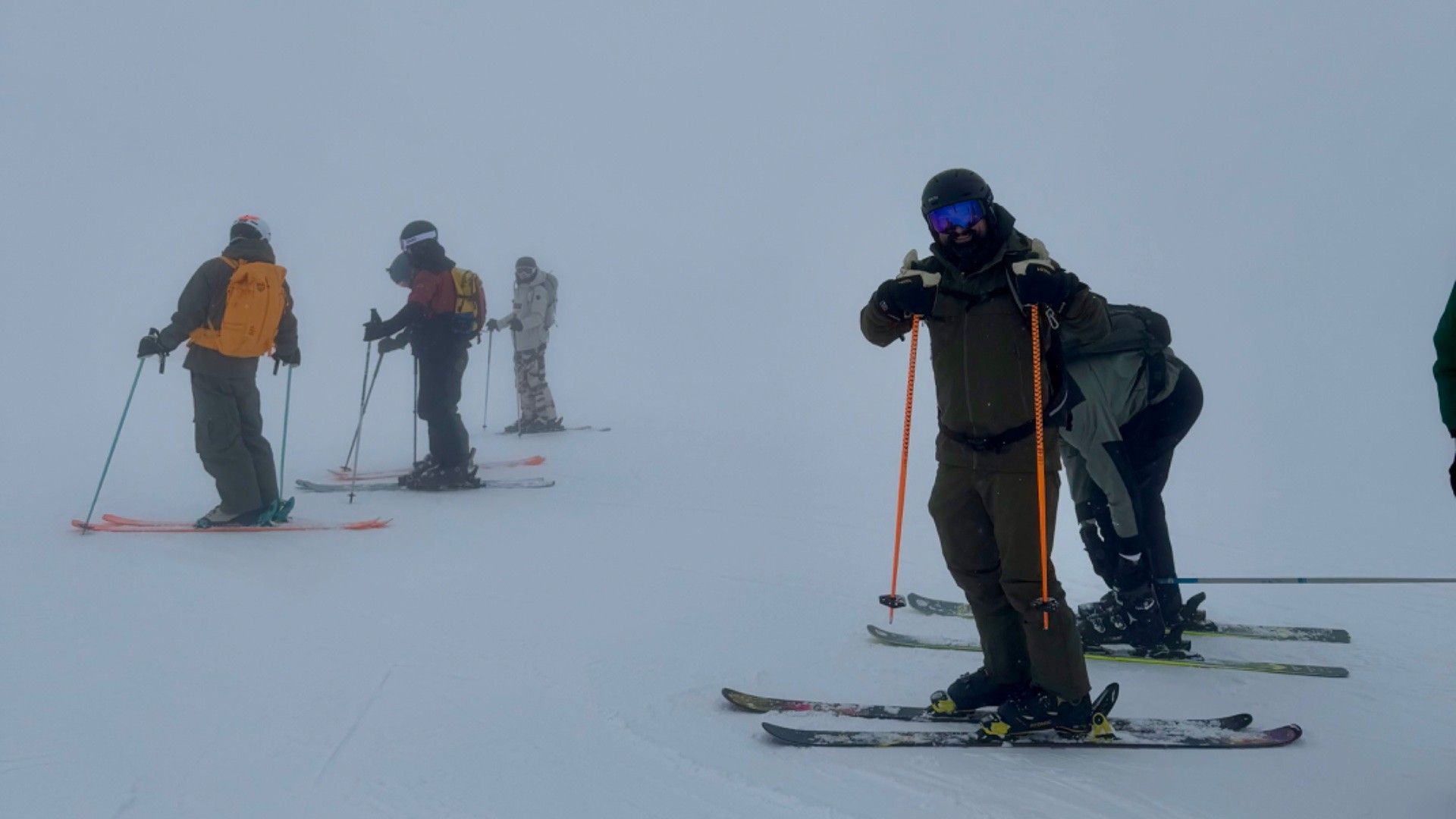 A group of skiers of varying abilities pause together on a snowy slope in mist